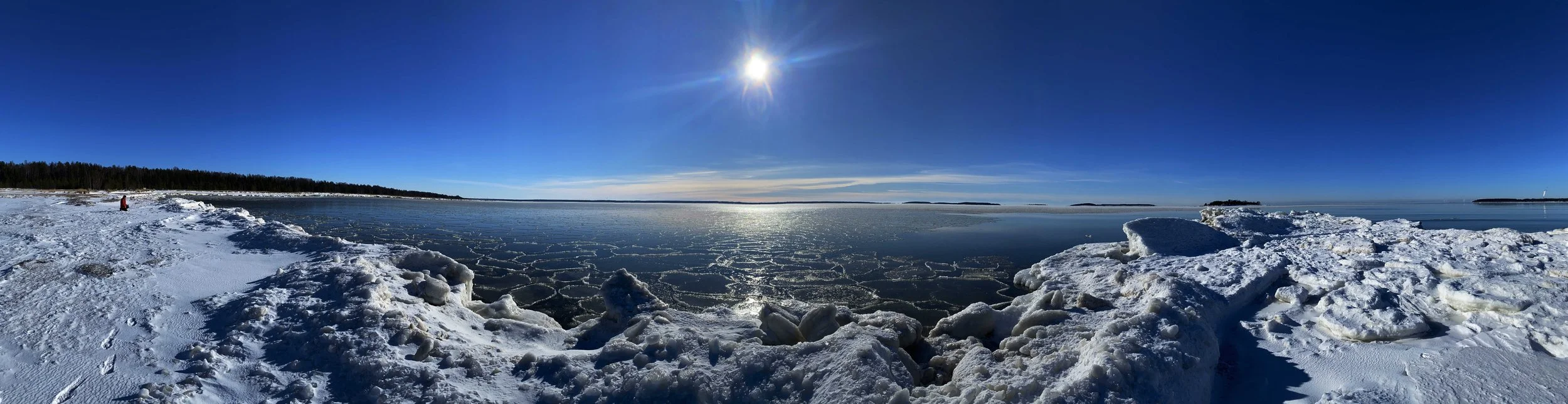 A snowy landscape with a frozen body of water under a clear blue sky and the sun shining brightly. Snow and ice cover the ground, with a person sitting on the snow in the distance
