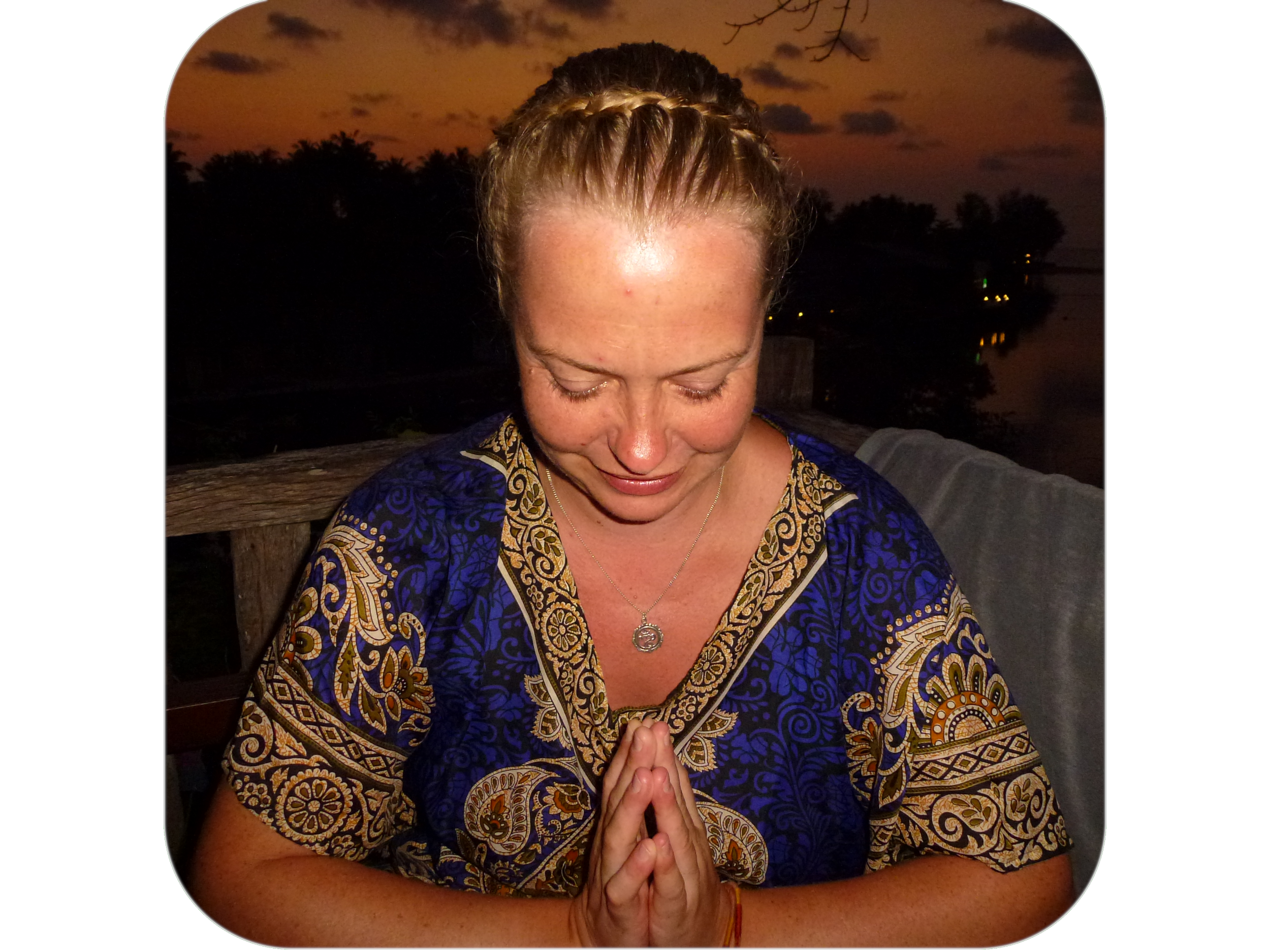 A woman with blonde hair in a braided updo, wearing a colorful patterned top and a necklace, is bowing her head with her hands pressed together in a prayer or meditative gesture, outdoors during sunset with a river and trees in the background.