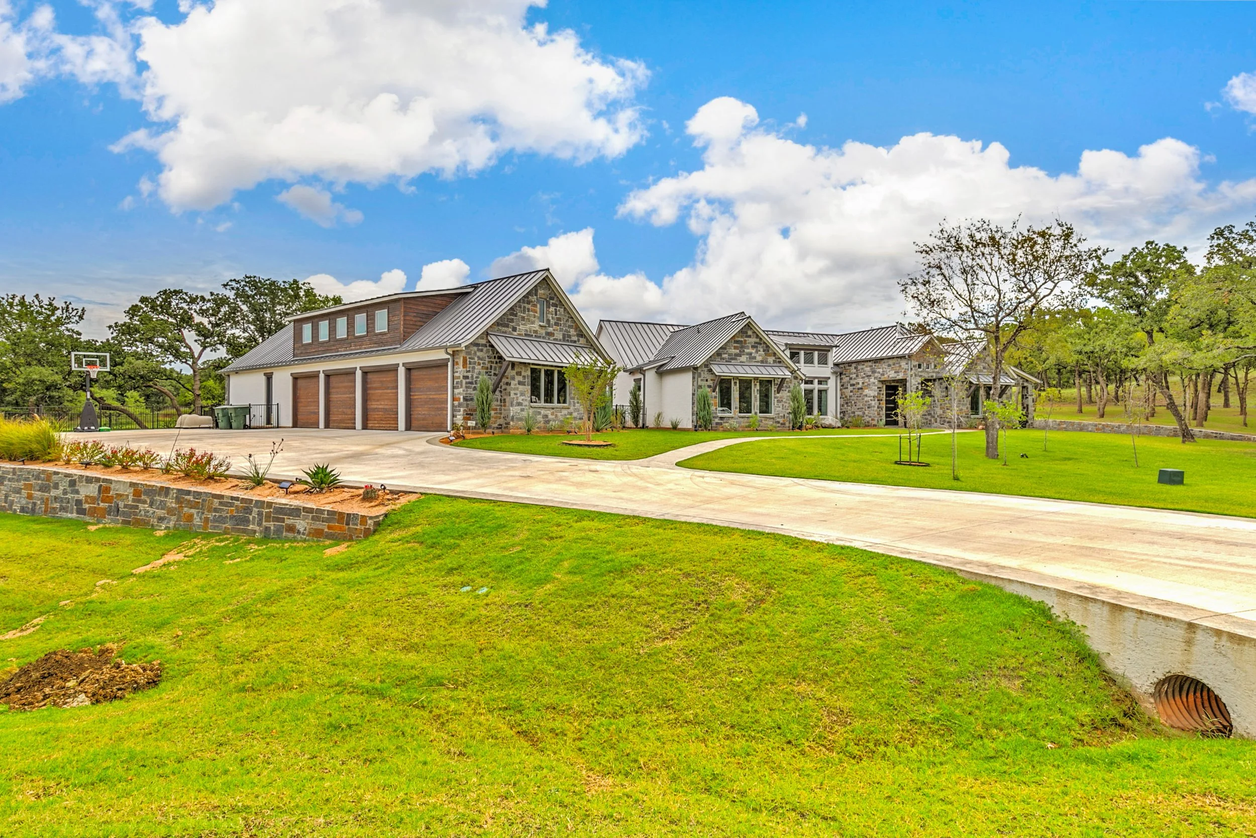 A modern house with stone and wood exterior, metal roof, and a driveway, set against a green lawn and trees under a partly cloudy sky.