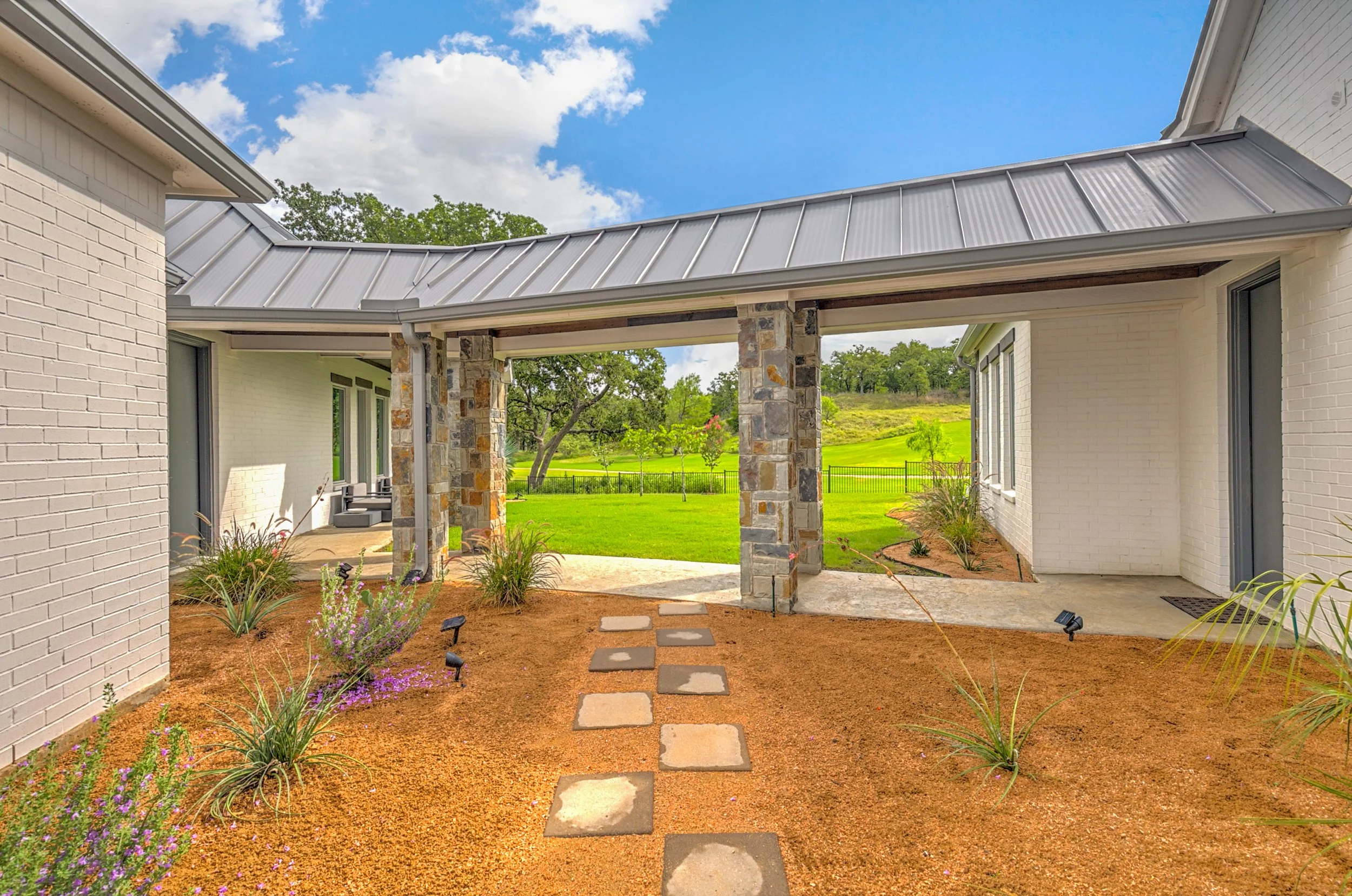 View of a backyard patio with a stone and brick column, stepping stones on mulch, and a backdrop of a green lawn, trees, and a partly cloudy sky.