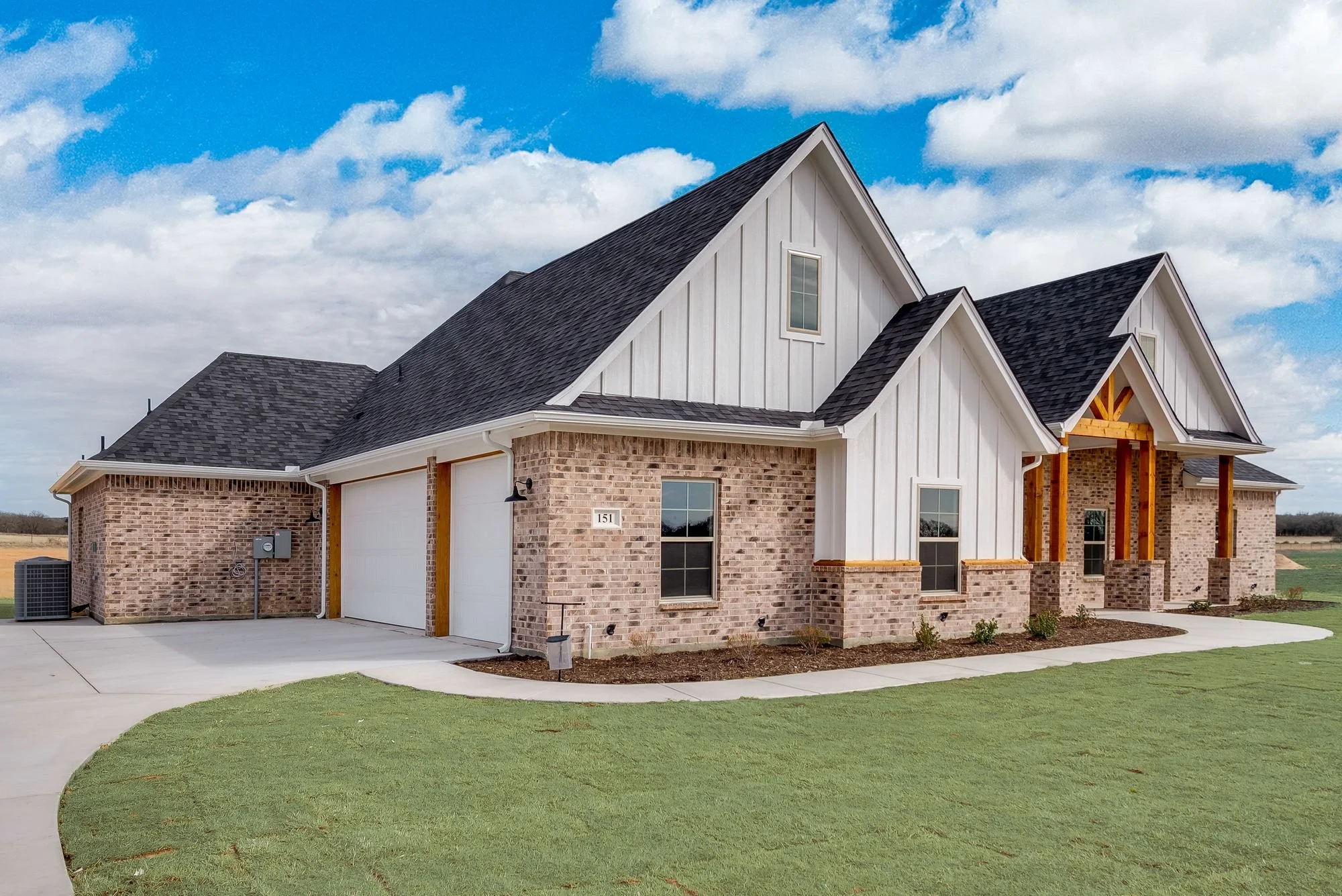 Newly built two-story house with a brick and white siding exterior, two garage doors, multiple windows, and a front porch under construction. The house is situated on a well-maintained lawn with a curved sidewalk.