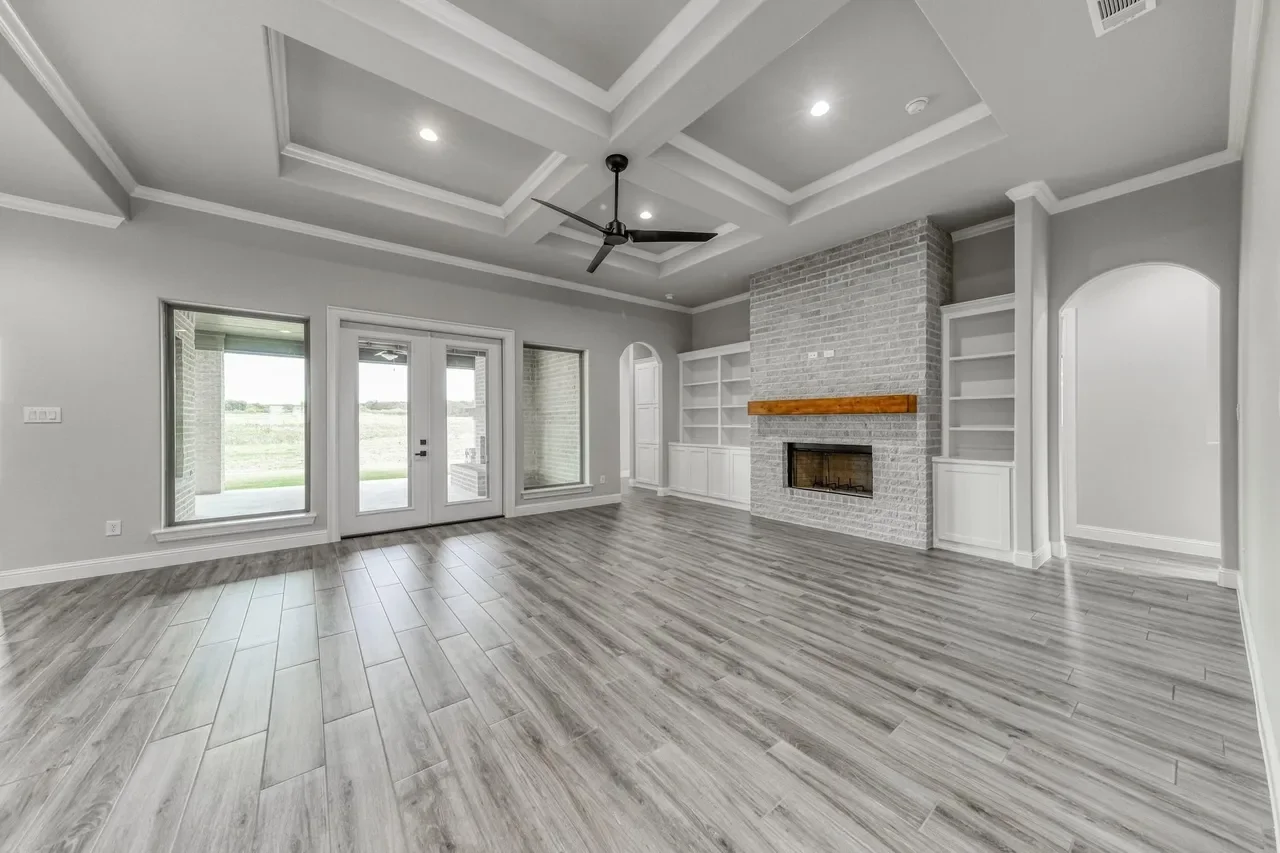 Empty living room with gray wood floors, a white brick fireplace with a wooden mantel, built-in white shelves on either side of the fireplace, large windows, glass double doors leading outside, and a coffered ceiling with a ceiling fan.