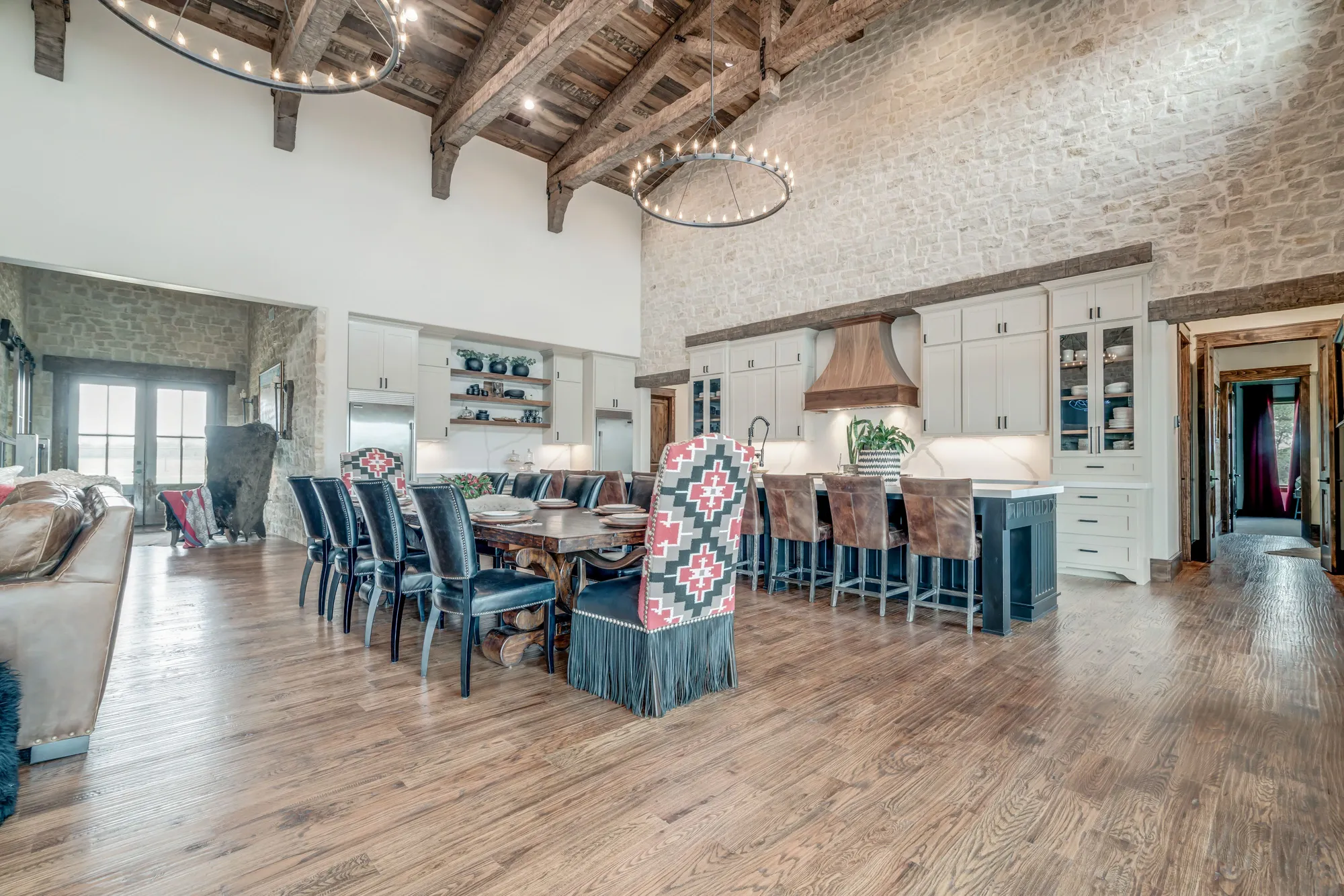 Open-concept living and dining area with a high vaulted ceiling featuring exposed wooden beams and chandeliers. A large wooden dining table is surrounded by chairs, some with patterned upholstery. The space has a rustic aesthetic with brick accents, white cabinets, and wooden floors.