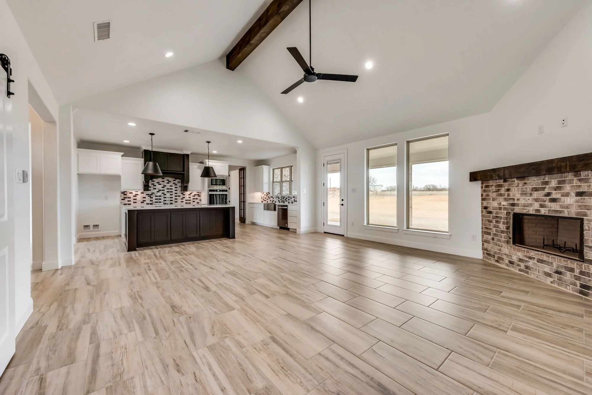 Open-concept living room and kitchen with wood-look tile flooring, a brick fireplace, large windows, ceiling fan, and modern kitchen cabinetry and appliances.