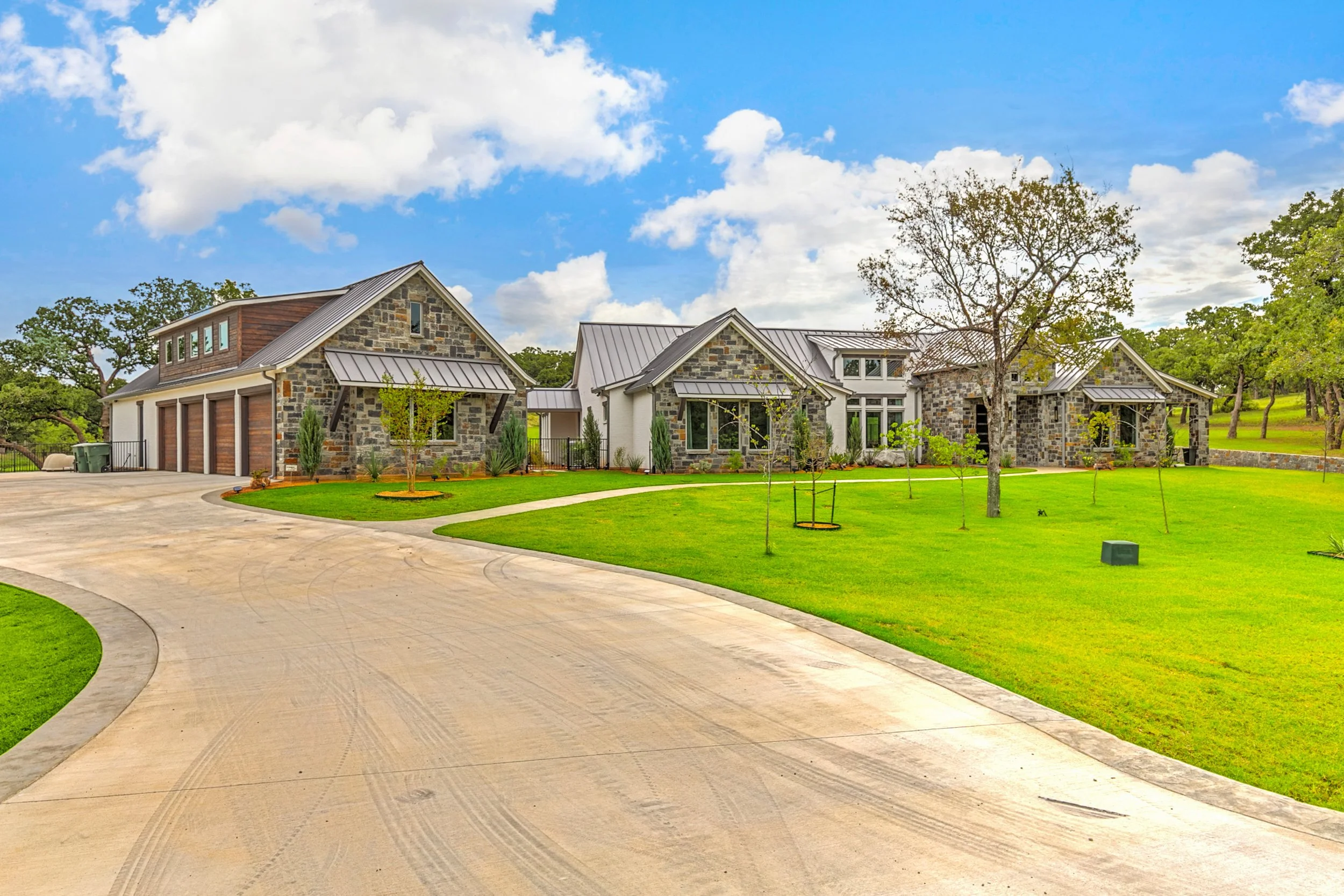 A modern house with stone and white siding, multiple gabled roofs with metal roofing, a three-car garage, paved driveway, surrounded by green lawn and trees, under a partly cloudy sky.