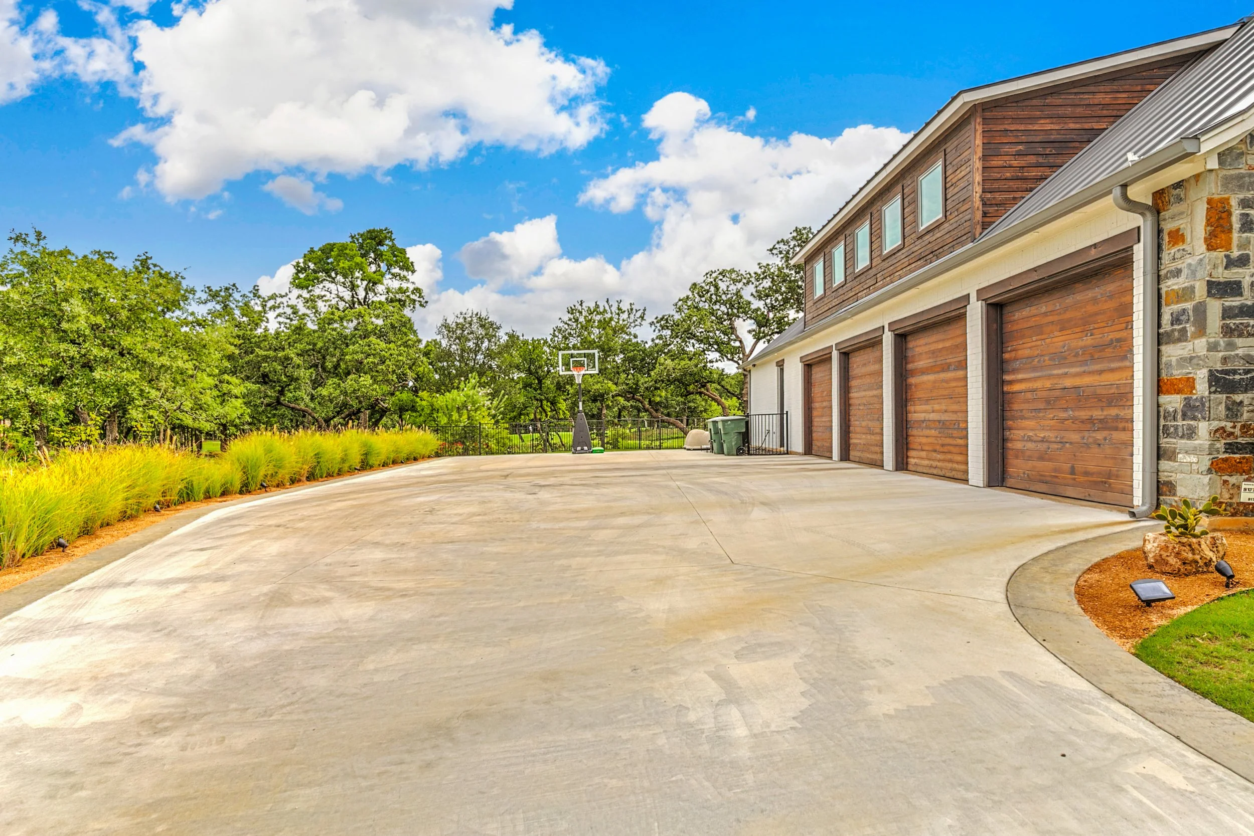 A modern house with a wooden and stone exterior, a multi-car garage, a concrete driveway, a backyard basketball hoop, surrounding greenery, and a blue sky with clouds.