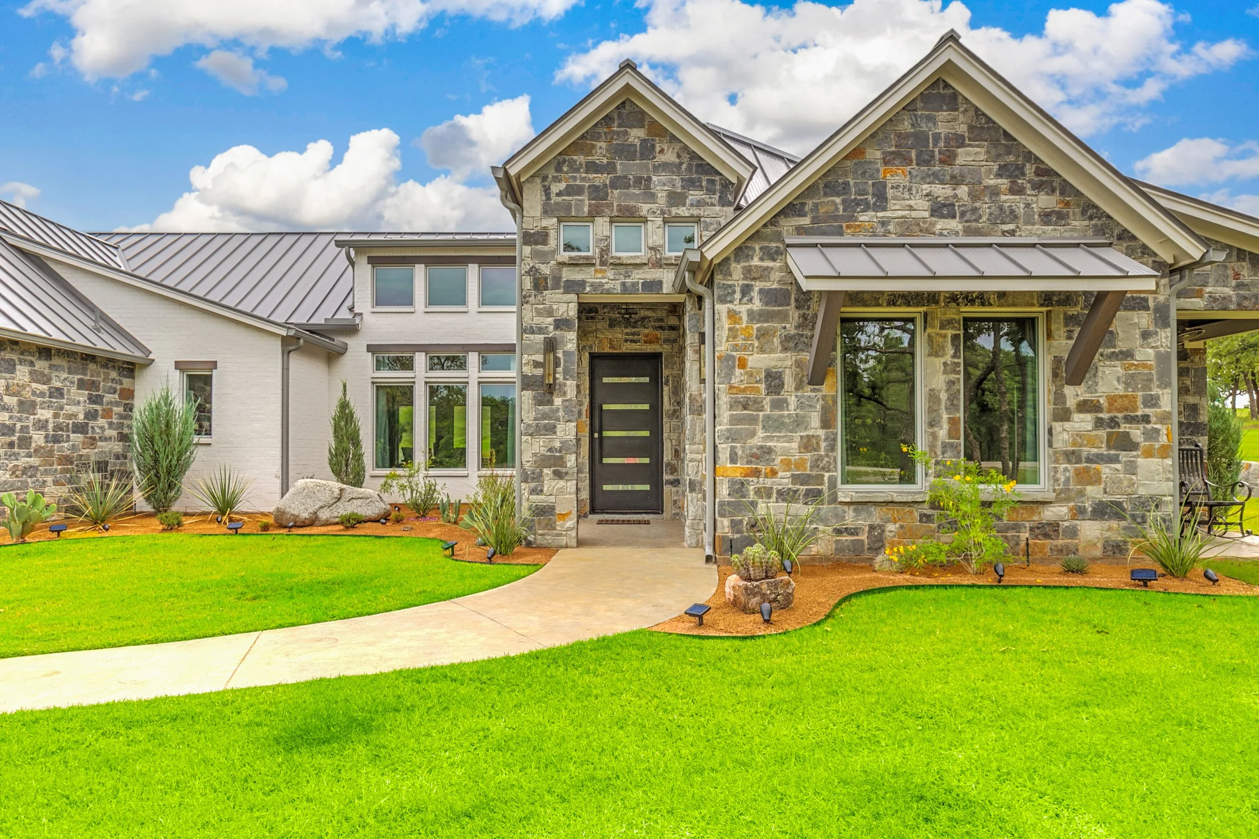 Front view of a modern house with a stone facade, large windows, a black front door, and a well-maintained green lawn under a partly cloudy sky.
