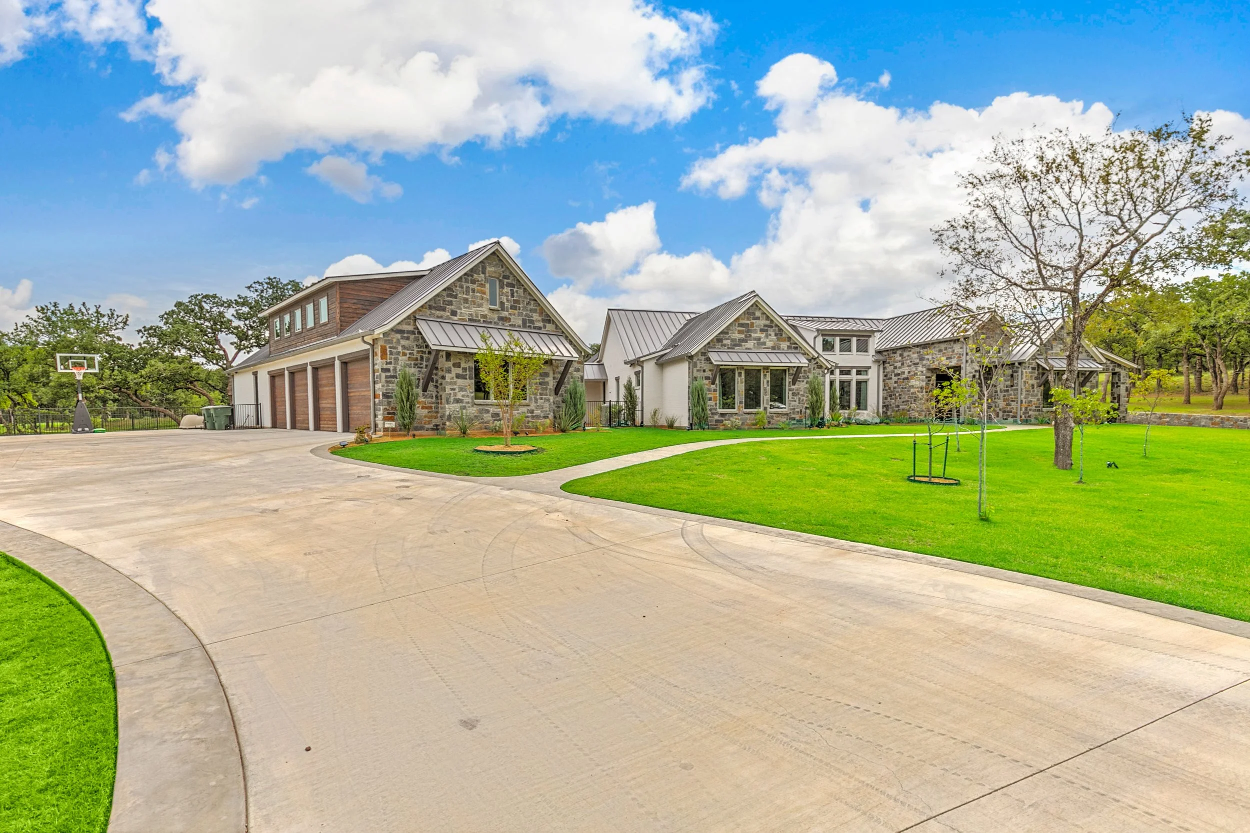 A modern house with stone and wood exterior, multiple gabled roofs, surrounded by a green lawn, young trees, and a driveway on a clear day with a blue sky and white clouds.