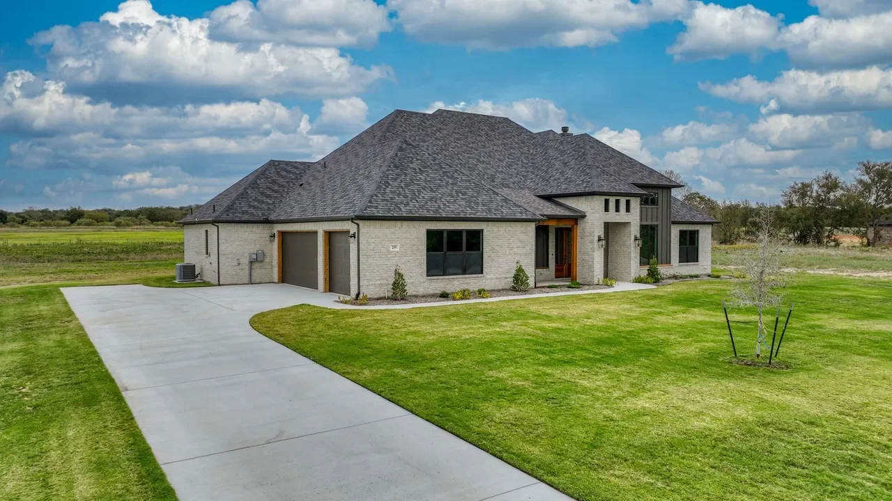 Newly built house with a gray roof, gray brick walls, and a front yard with a curved concrete driveway, green lawn, young tree, and small shrubs.