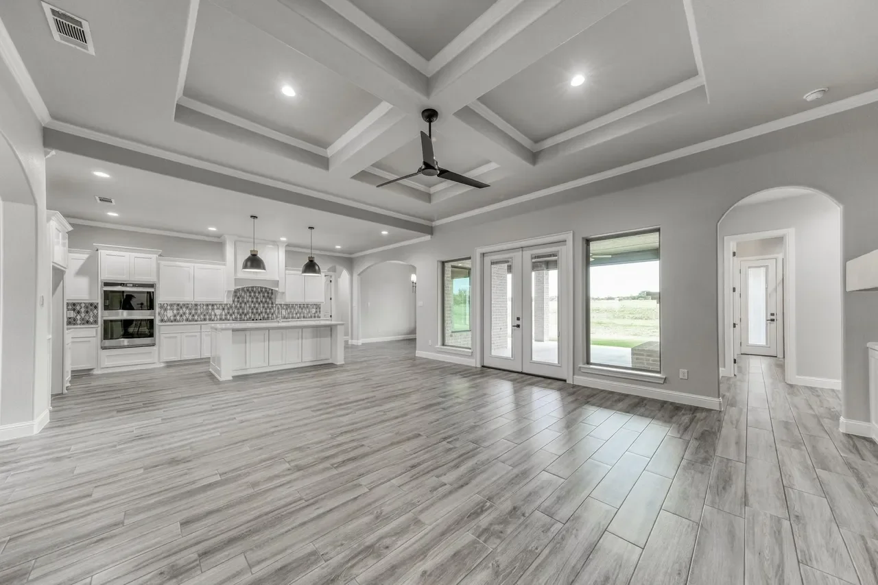 Empty open-concept living area with gray wood floors, white walls, large windows, and a kitchen with white cabinets and patterned backsplash.