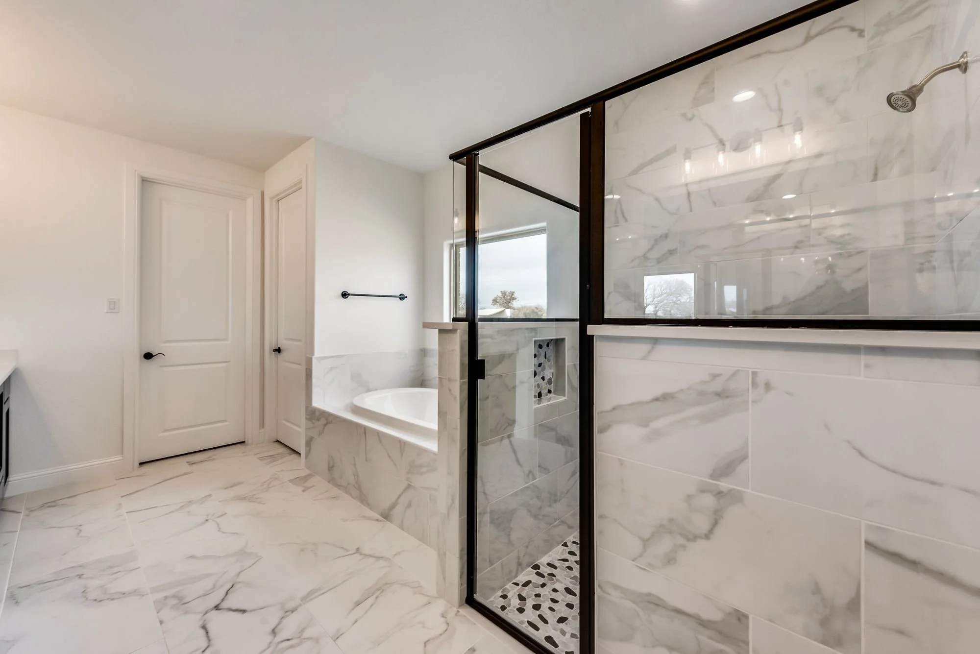 Modern bathroom featuring a marble-tiled floor and walls, a bathtub with marble surround, and a glass-enclosed shower with marble and pebble tile accents. There are two white doors and a window showing a tree outside.