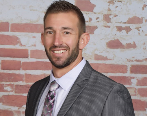 A smiling man with short dark hair and a beard, wearing a gray suit, white shirt, and a plaid tie, standing in front of a red brick wall.