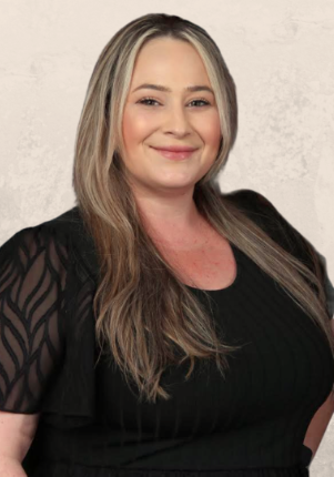 A woman with long blonde hair smiling, wearing a black top and black patterned short-sleeve jacket, standing against a light-colored textured wall.