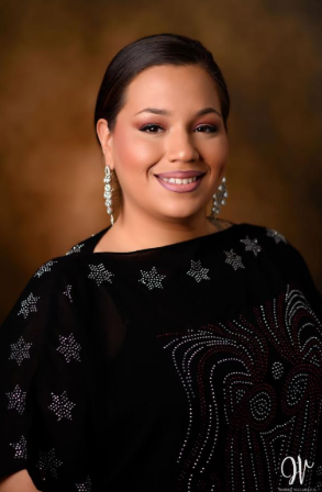 A woman with dark hair styled back, wearing makeup, diamond earrings, and a black dress with silver star and swirl patterns, smiling against a brown background.