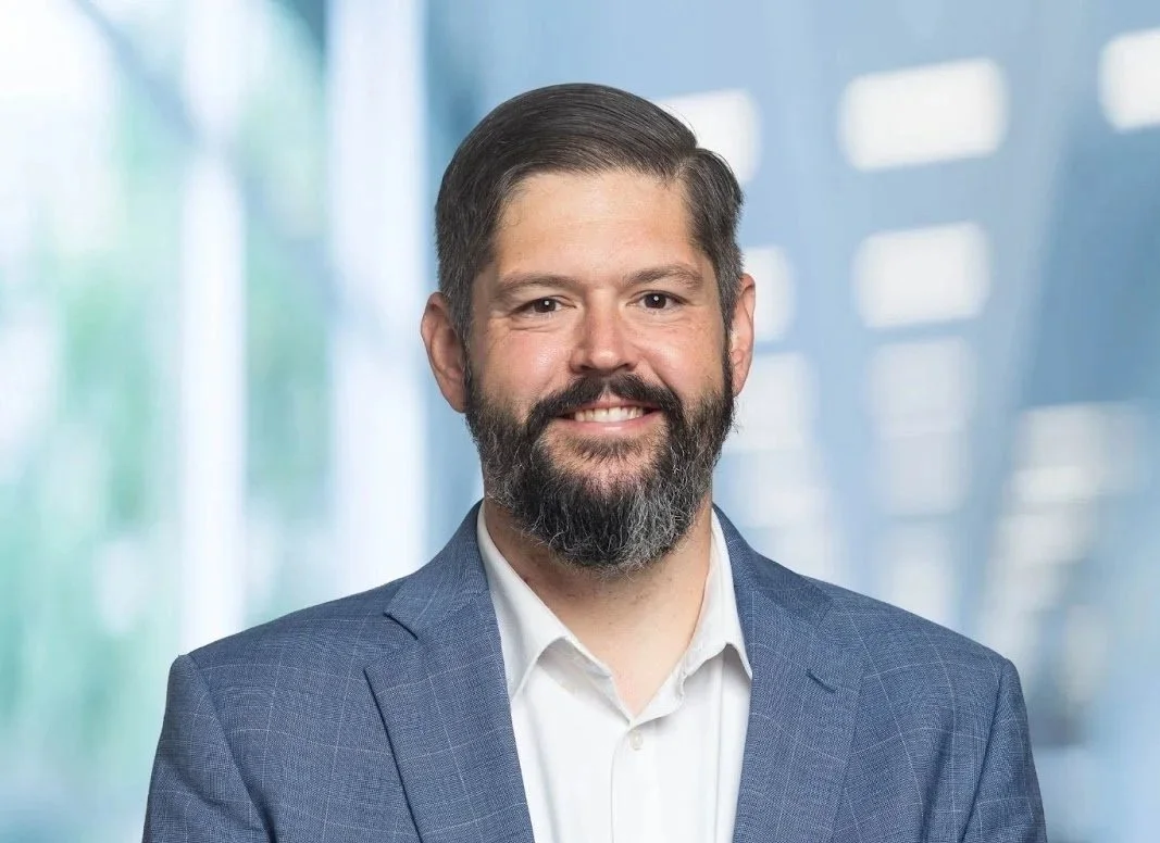 Professional man with a beard and short dark hair, wearing a blue suit jacket and white shirt, standing in an office setting with blurred background.