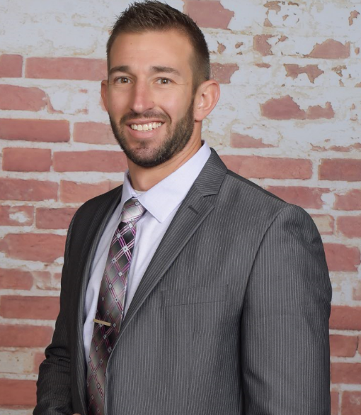 Portrait of a smiling man in a gray suit, white shirt, and patterned tie standing in front of a brick wall background.