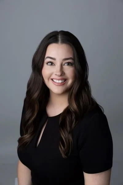 A young woman with long dark hair, smiling and wearing a black top, against a plain gray background.