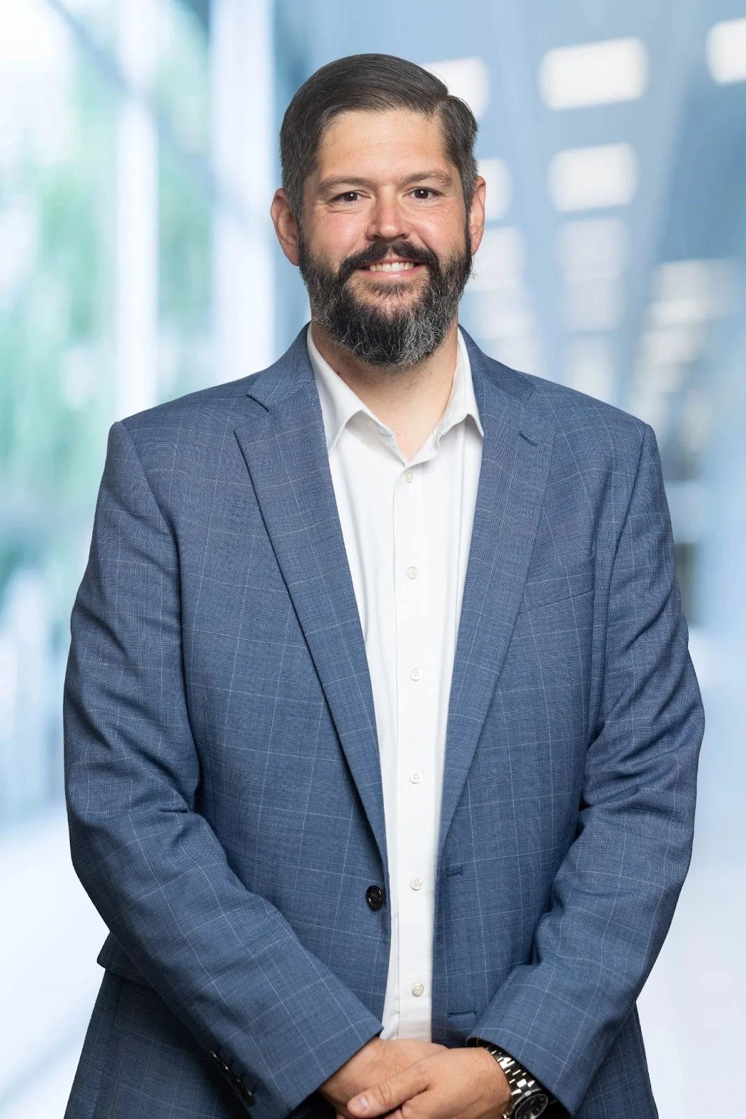 A man in a blue plaid blazer and white shirt smiling, standing in a bright, modern office corridor.