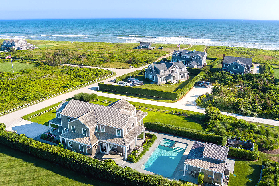 Aerial view of beach houses along the shoreline, with a house featuring a swimming pool in the foreground and the ocean in the background.