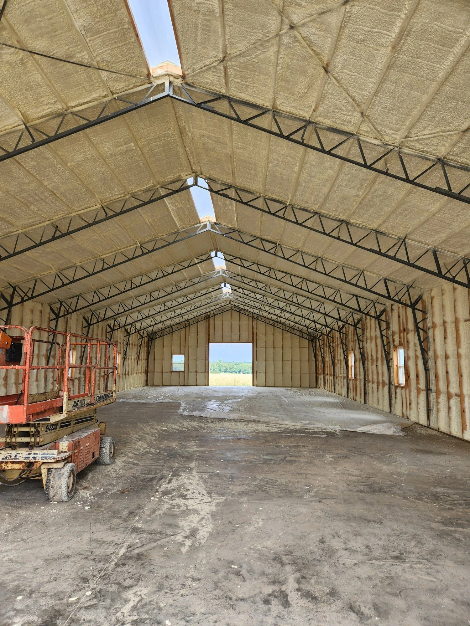 Interior of a large, unfinished metal building under construction with insulation on the walls and roof, a scissor lift on the left, and an open doorway in the back showing a view of the outdoors.