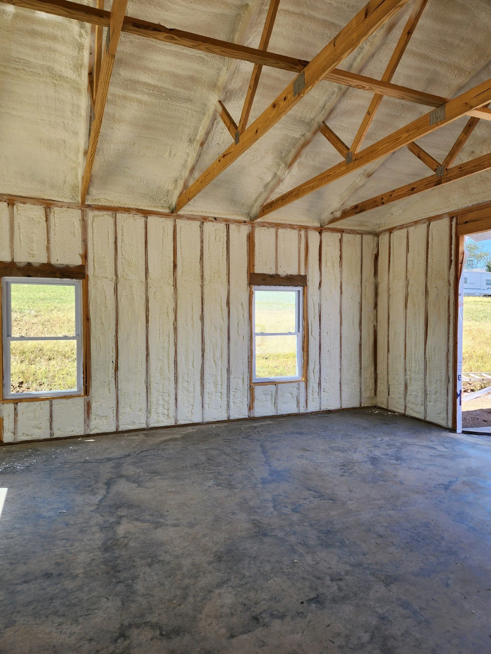 Interior view of a house under construction with unfinished walls and exposed ceiling framing, showing two windows and a doorway.