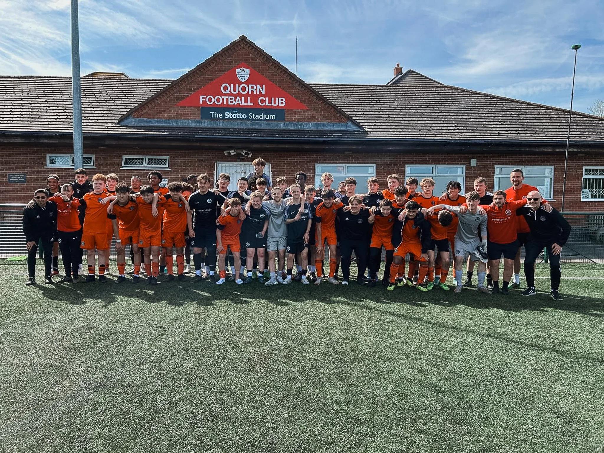 Group of young soccer players and coaches standing on a soccer field in front of a building with a sign that reads 'Quorn Football Club' and 'The Stotto Stadium'.