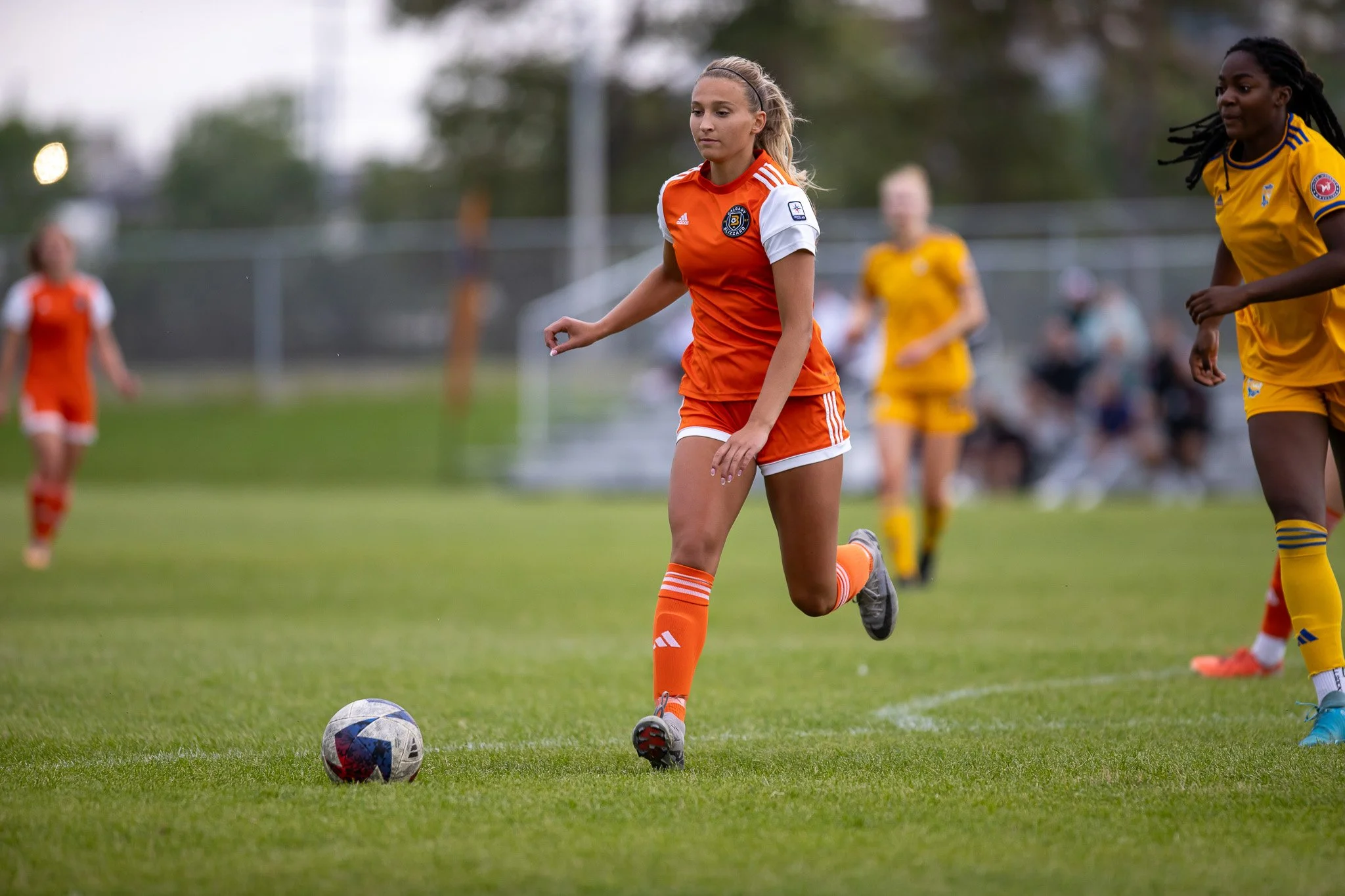 A young female soccer player in an orange uniform controls the ball on a soccer field during a game, with other players visible in the background.
