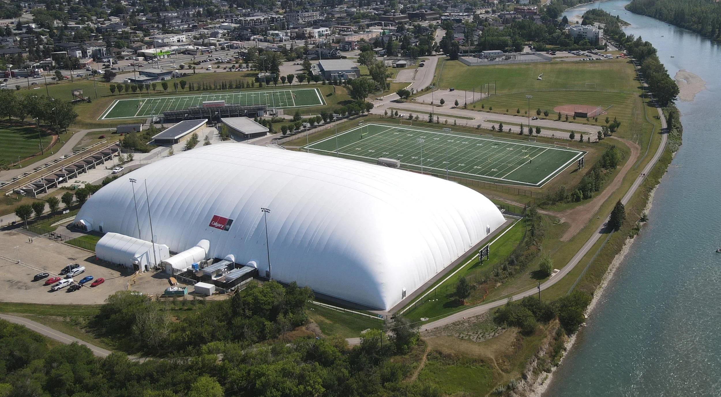 An aerial view of a sports complex with a large white dome, multiple football fields, and a river on the right side.