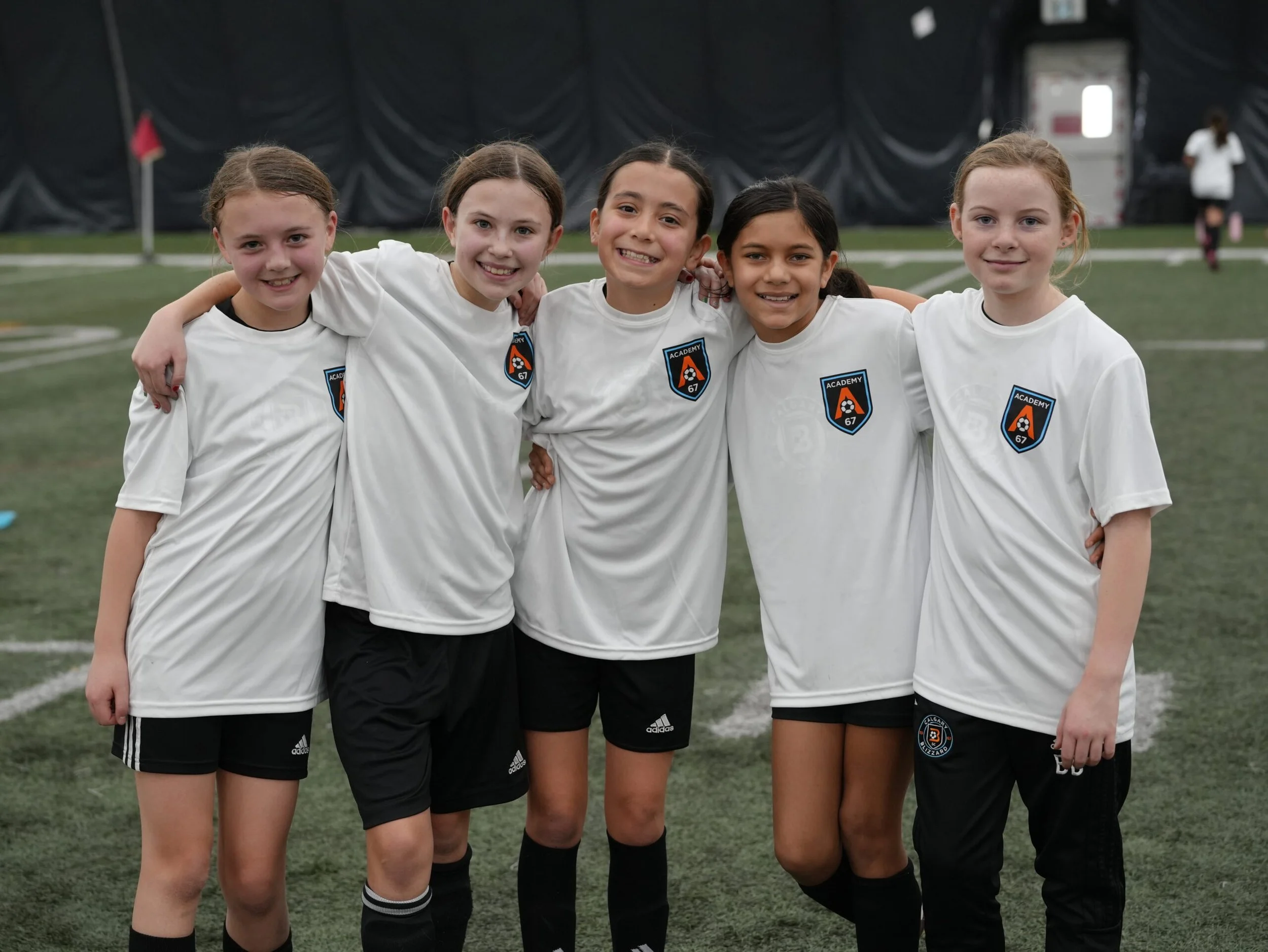 Five young girls standing together on an indoor soccer field, wearing white soccer jerseys with a blue and orange team logo. They are smiling and have their arms around each other.
