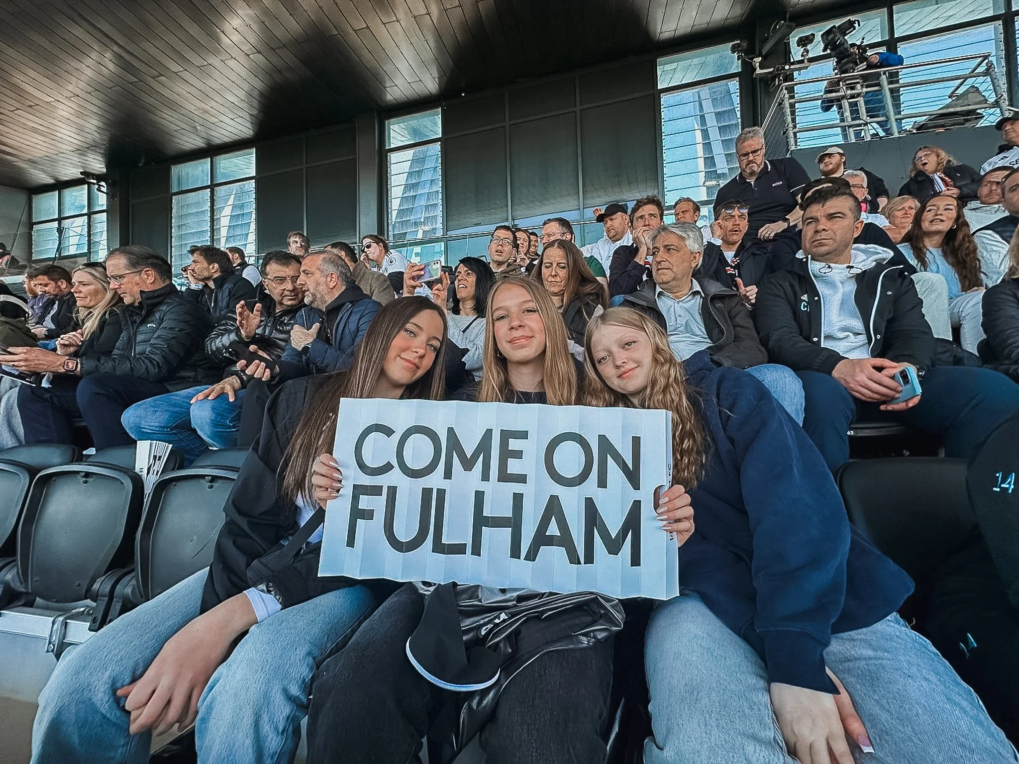 Three young girls sitting in stadium seats holding a sign that says "Come on Fulham" during a sports event with fans in the background.