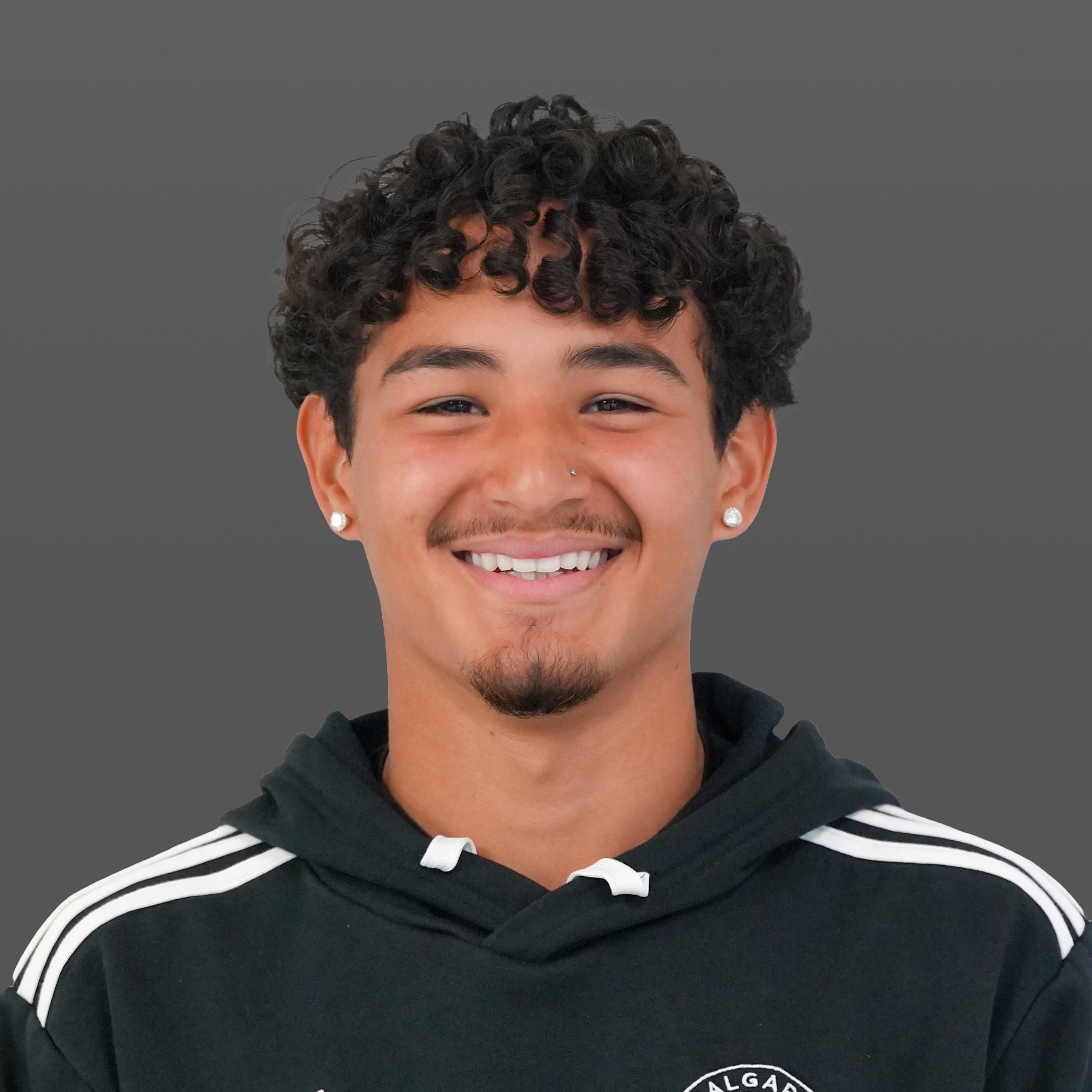 A young man with curly dark hair, tan skin, nose piercing, earrings, and a goatee, smiling at the camera while wearing a black hoodie with a teal Adidas logo, standing on an illuminated sports field at night.