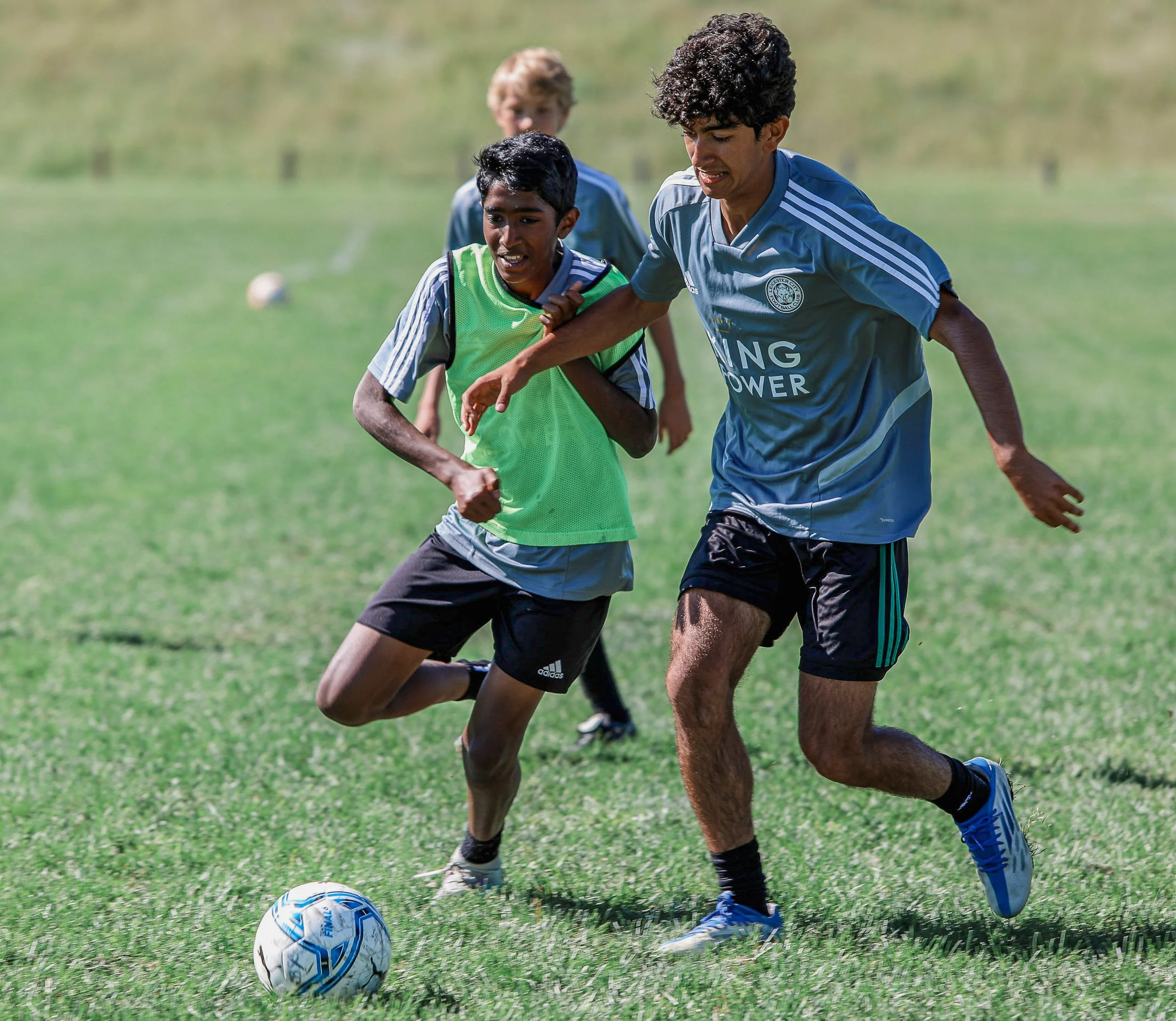 Two soccer players, a young boy and an older male, are running after a soccer ball on the field during a game. The boy is wearing a green vest, and the adult is wearing a blue jersey. They are focused and moving quickly, with other players visible in