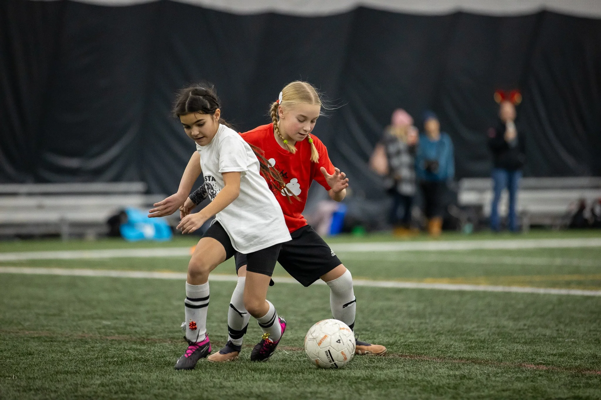 Two young girls playing soccer indoors, trying to gain control of the ball. One girl is wearing a white shirt and black shorts, the other is wearing a red shirt and black shorts. In the background, several people are watching, some standing and some 