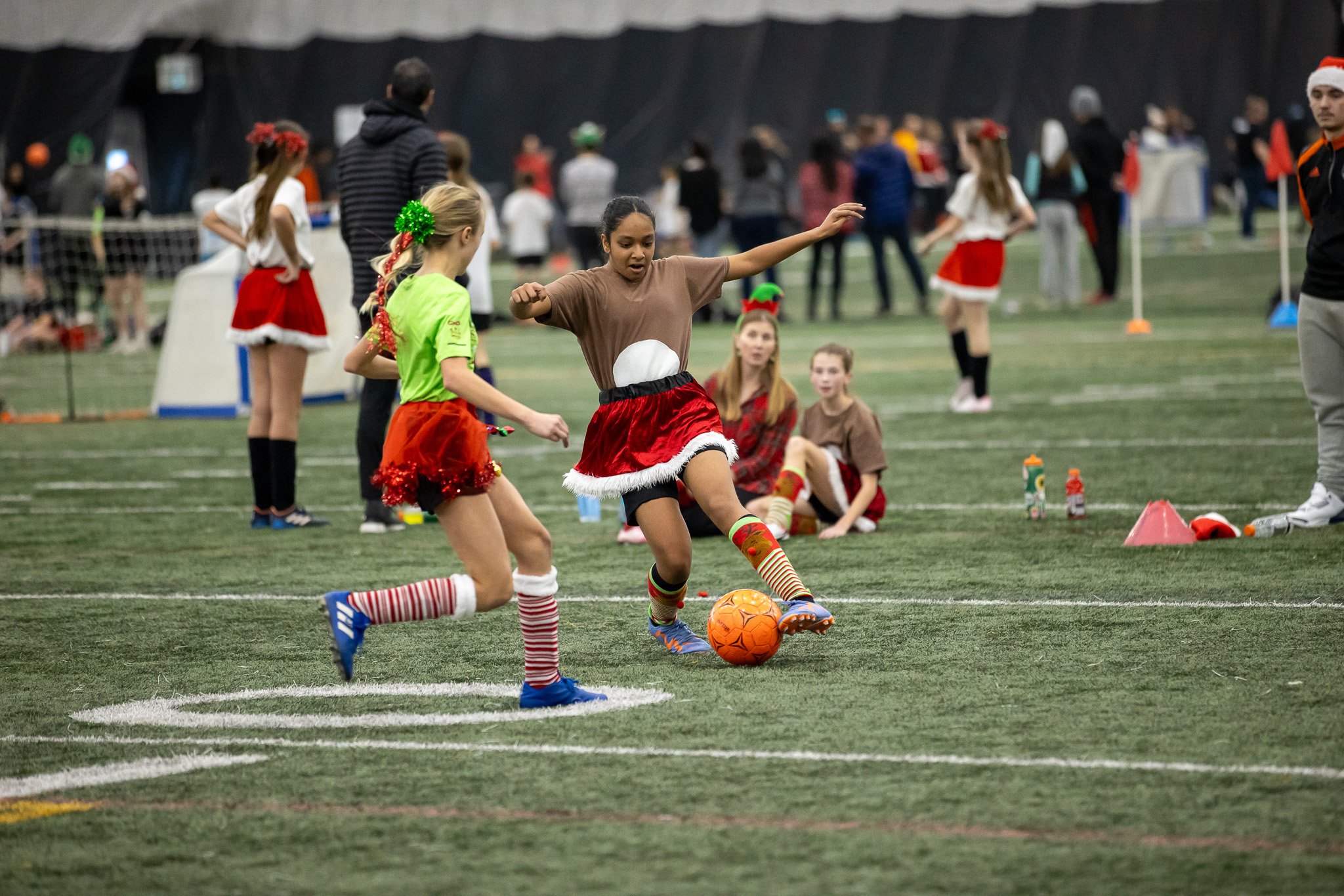 Young girls playing soccer on an indoor field during a Christmas-themed event, wearing festive outfits and holiday accessories.