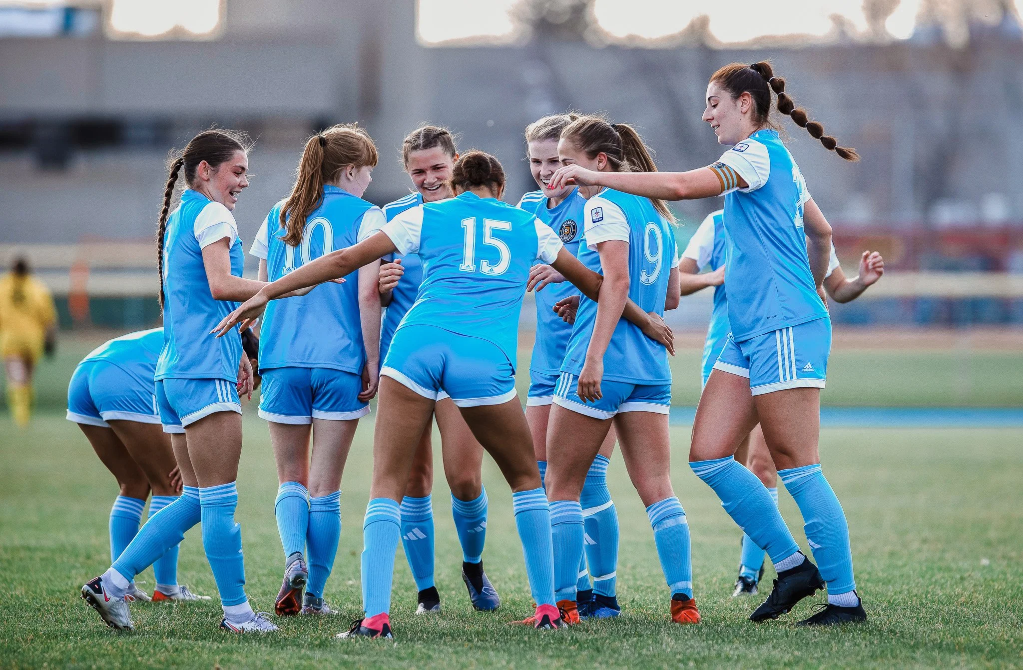 A group of female soccer players in blue and white uniforms celebrating together on a soccer field.