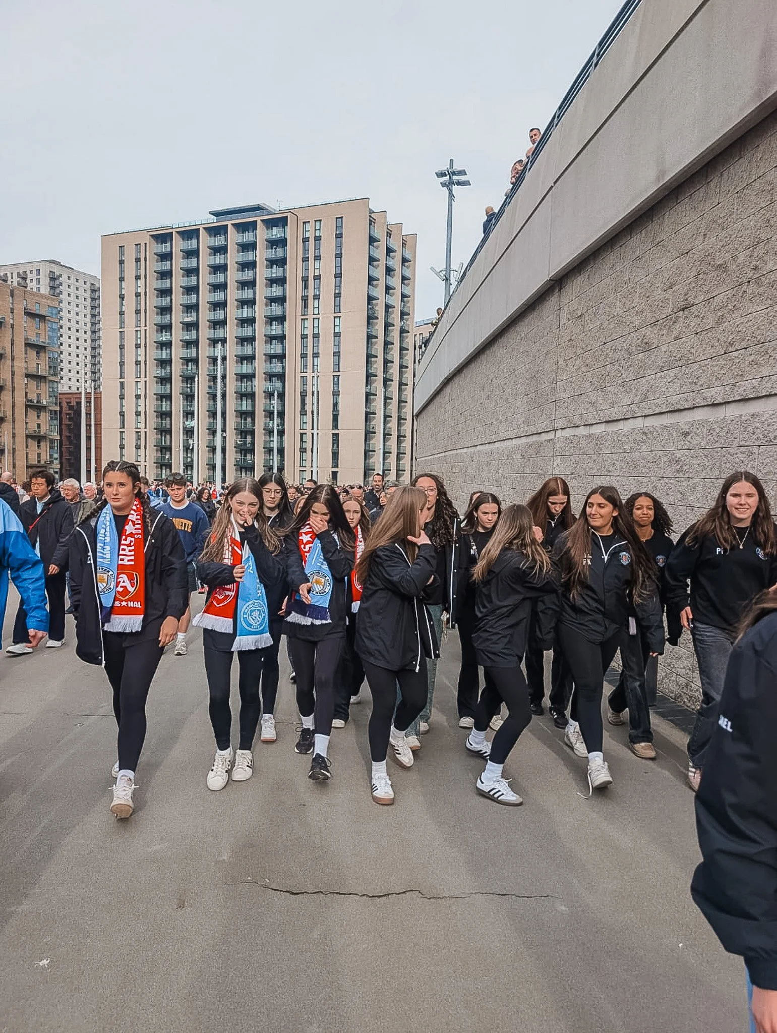 Group of young women walking outdoors near a large concrete wall, some wearing sports jackets and scarves, with high-rise buildings and a cloudy sky in the background.
