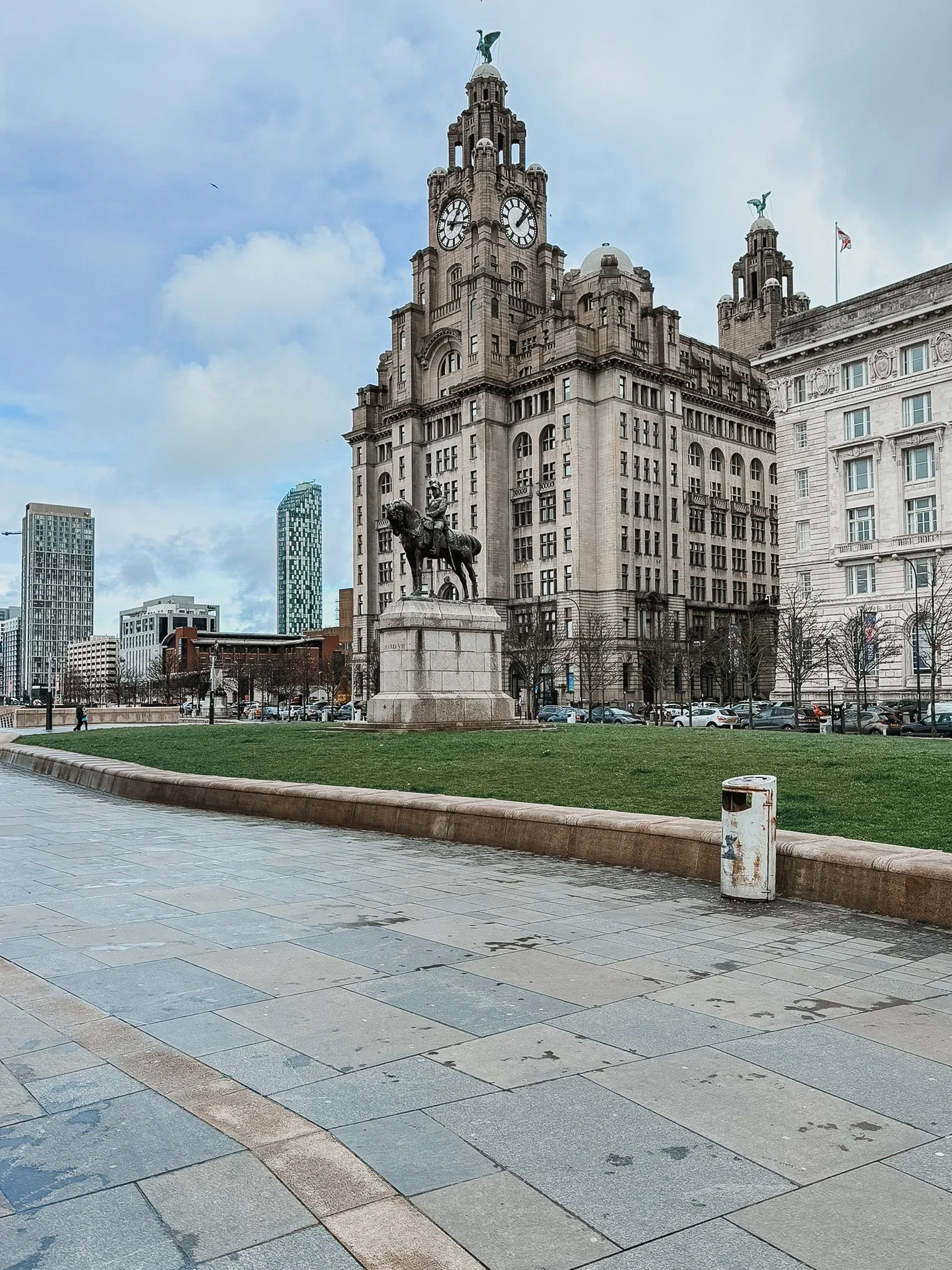 A historic clock tower building with a statue of a man on horseback in front, surrounded by modern skyscrapers, cars parked along the street, and a cloudy sky.