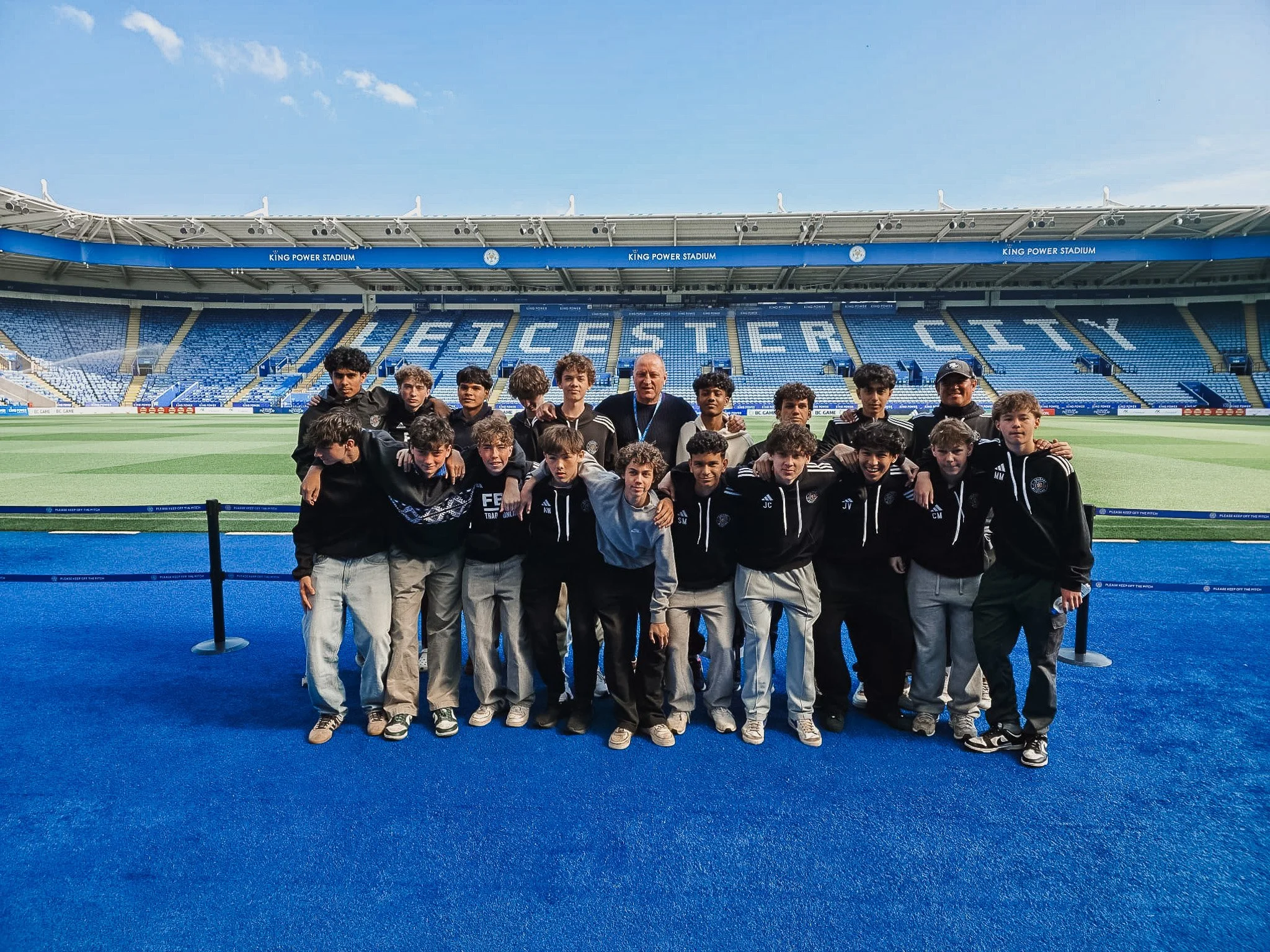 A group of young soccer players and two adults posing on the field at King Power Stadium in Leicester, UK, with empty blue seats and stadium infrastructure in the background.