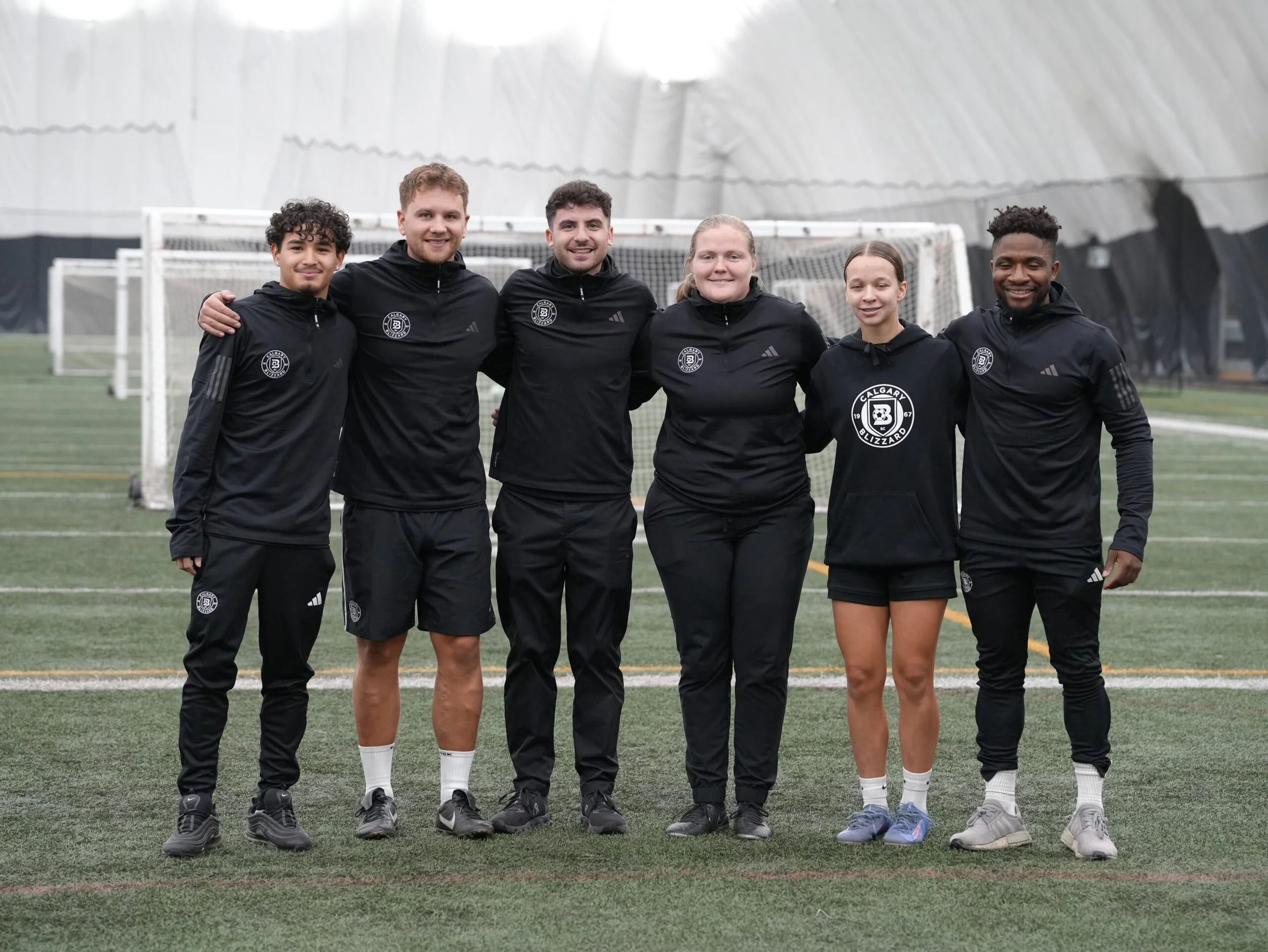 Group of six diverse soccer players standing together on a field inside an indoor sports dome, posing for a photo with arms around each other.