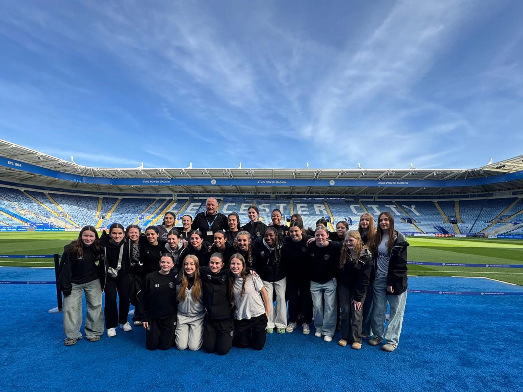 A group of young women and a coach standing together on a sports field inside Leicester City stadium, with empty blue seats and a bright blue sky in the background.