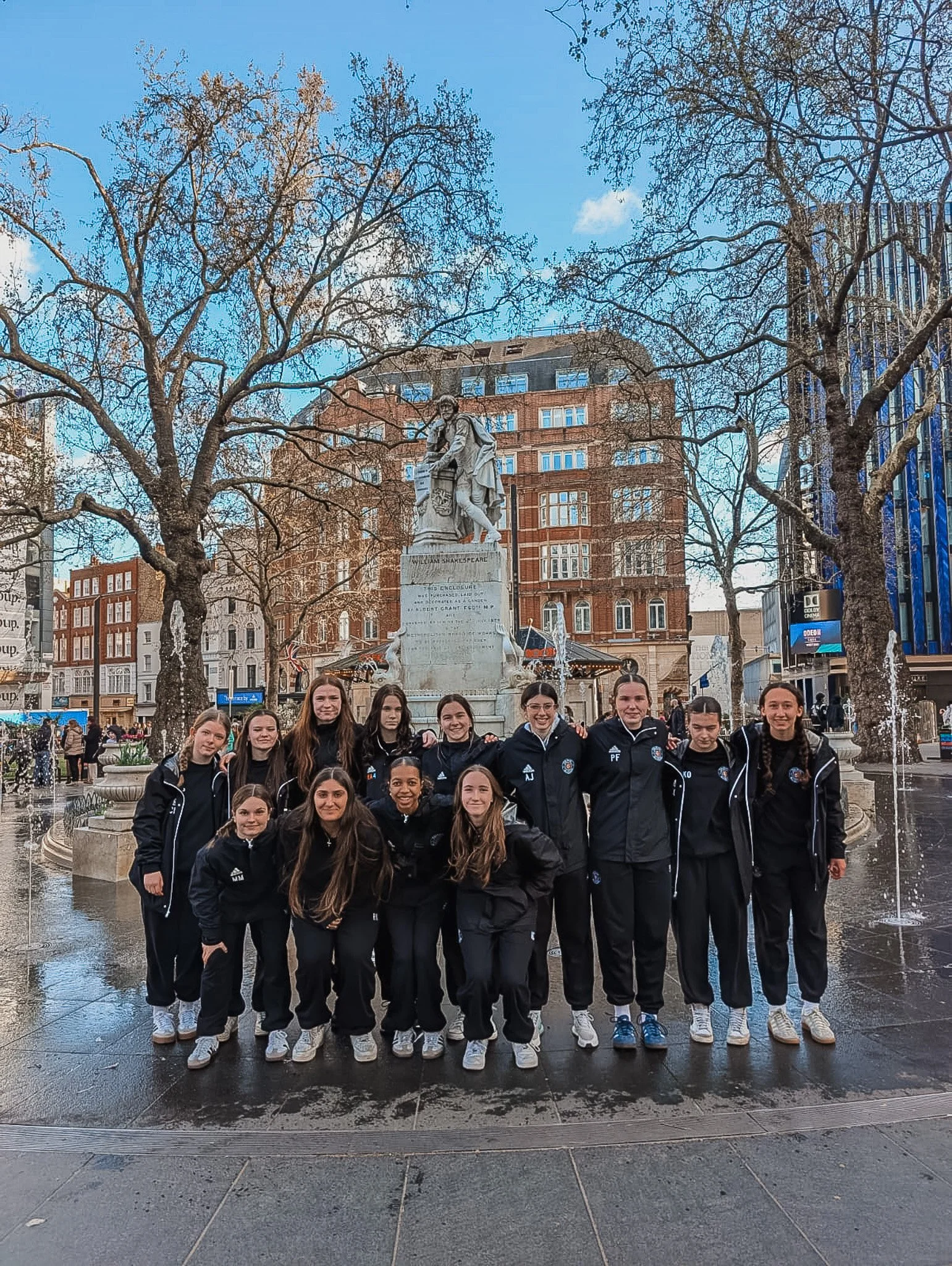 A group of girls in athletic jackets and pants standing in front of a fountain with a William Shakespeare statue in a city square, with trees, buildings, and a blue sky in the background.