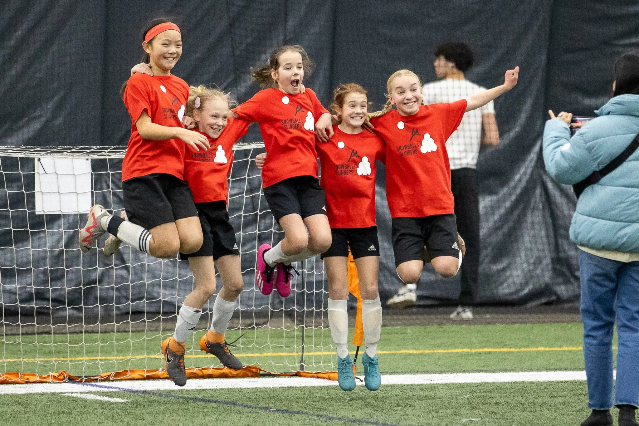 Five young girls in red jerseys and black shorts jumping in front of a soccer goal, smiling and celebrating during a game.
