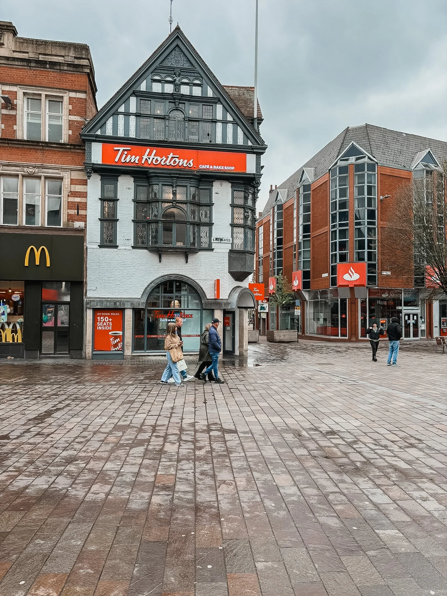 Street scene with pedestrians walking on wet cobblestone pavement. In the background, there is a building with a vintage design housing a Tim Hortons café and bake shop. To the left, a McDonald's restaurant is visible. To the right, there is a modern