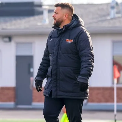 A man wearing a dark jacket with a hood and gloves standing outdoors near a building.