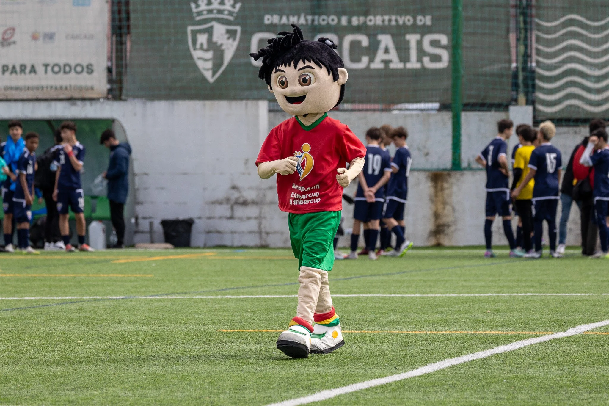 A person in a cartoon mascot costume running on a soccer field, with young players in uniforms standing in the background during a sports event.