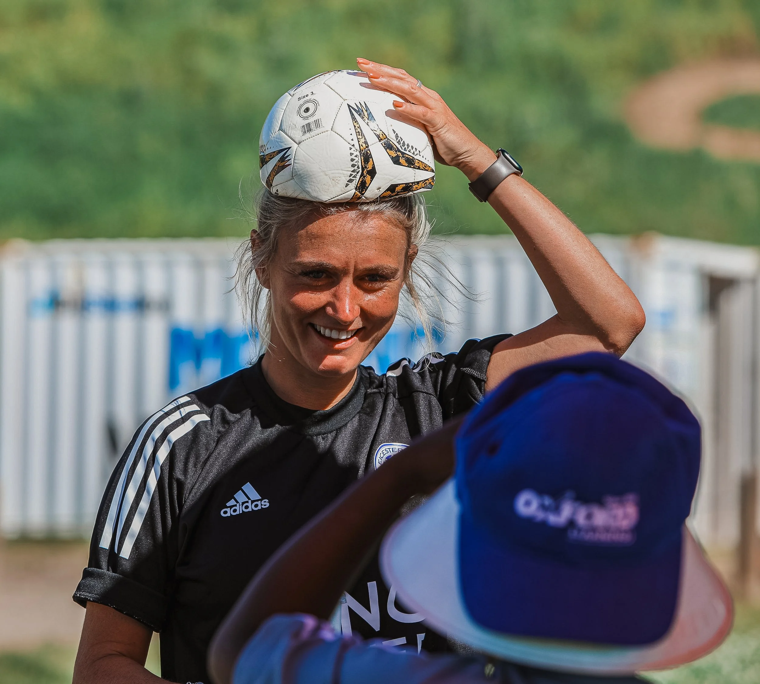 A woman smiling outdoors holding a soccer ball on her head, wearing a black Adidas sports shirt, with another person in a blue and white cap in front of her.