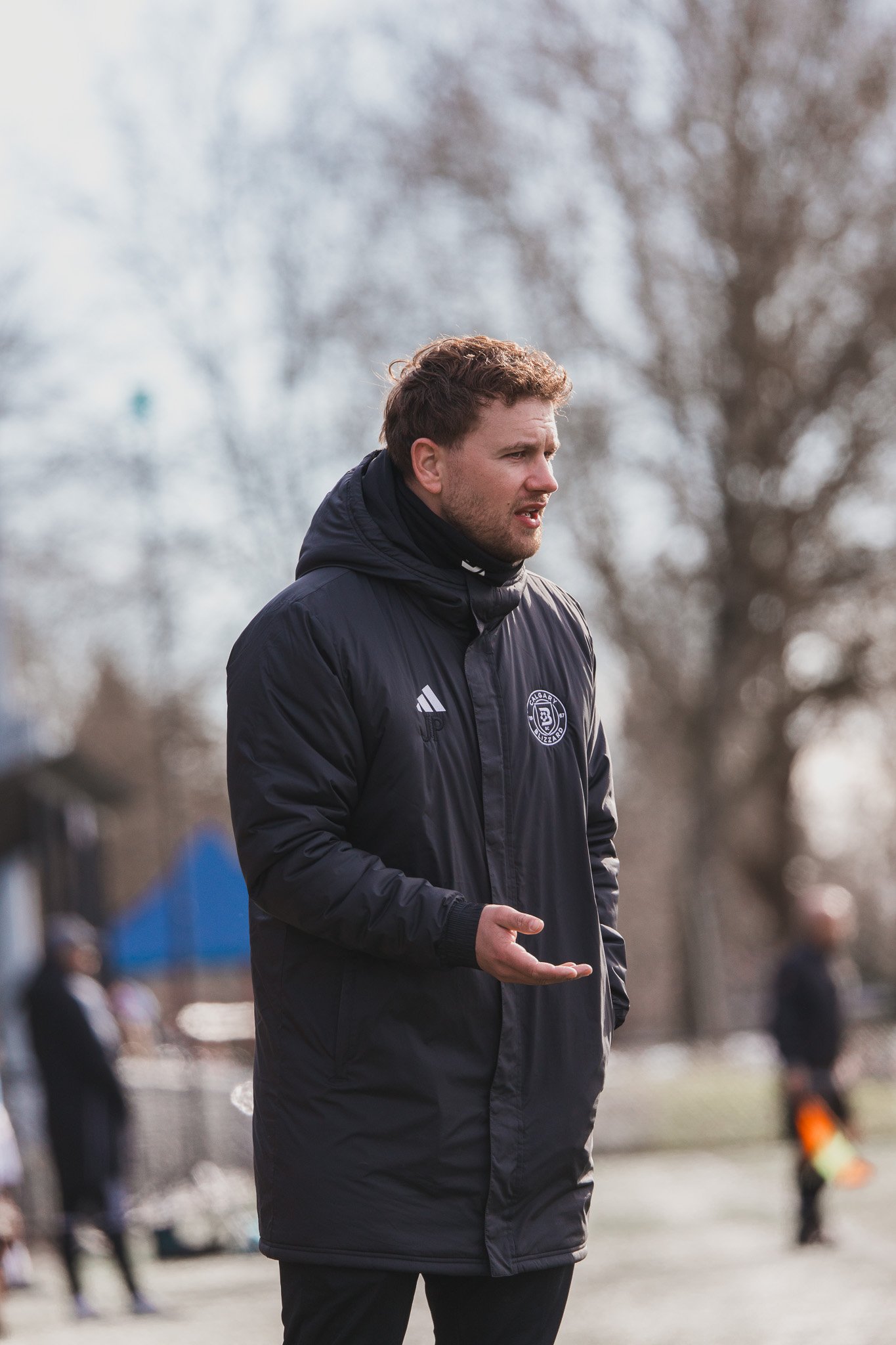 A man wearing a black jacket with a sports team logo, standing outdoors on a cloudy day, appears to be speaking or coaching.