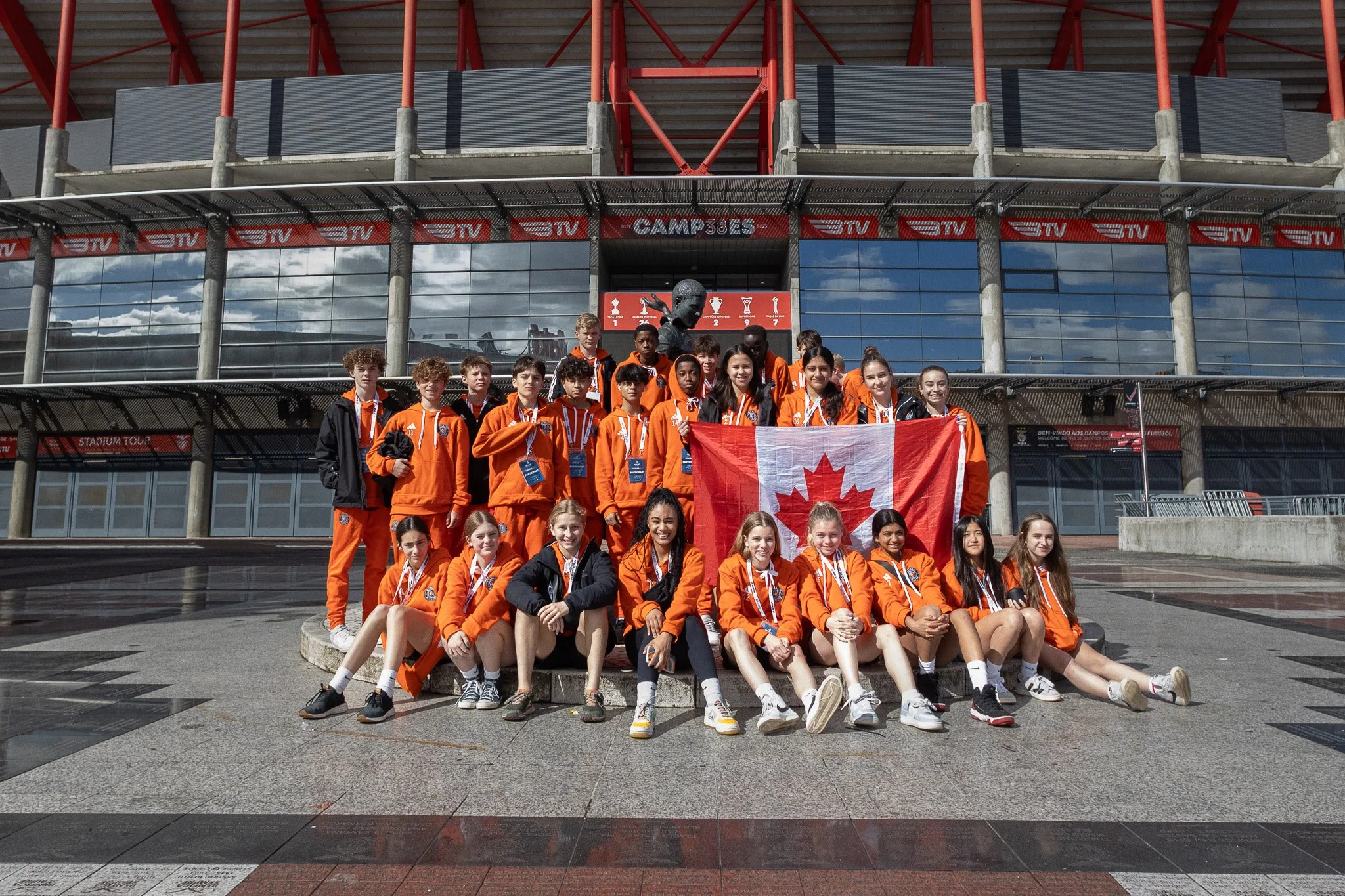 A group of young athletes in orange sportswear posing in front of a stadium, holding a Canadian flag.