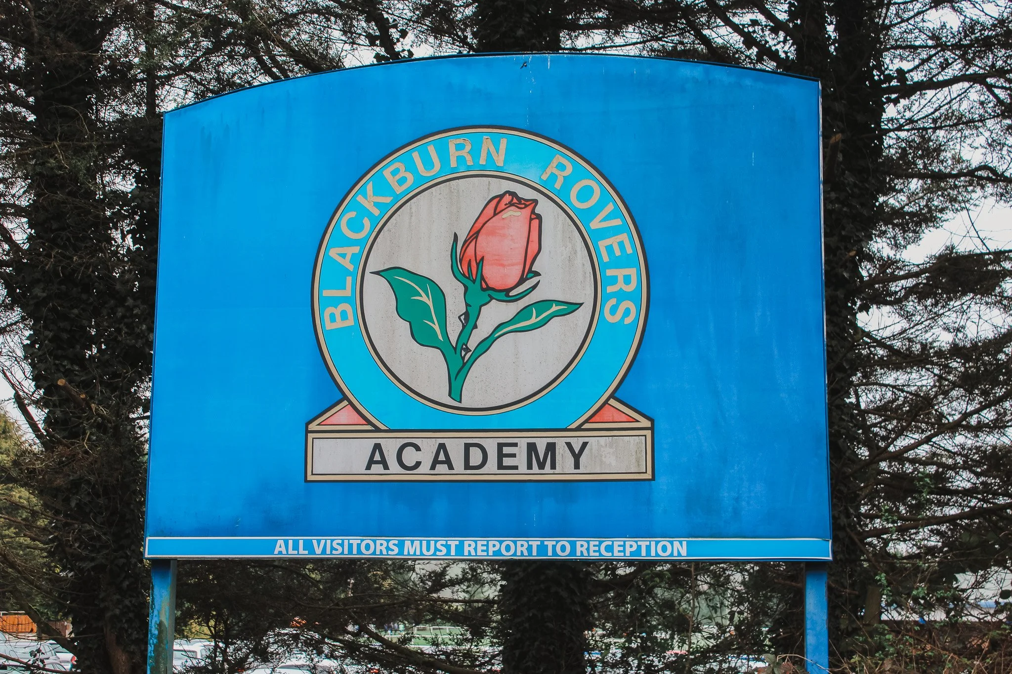 Blue sign for Blackburneers Rovers Academy with a rose emblem and a warning that all visitors must report to reception, positioned outdoors with trees in the background.