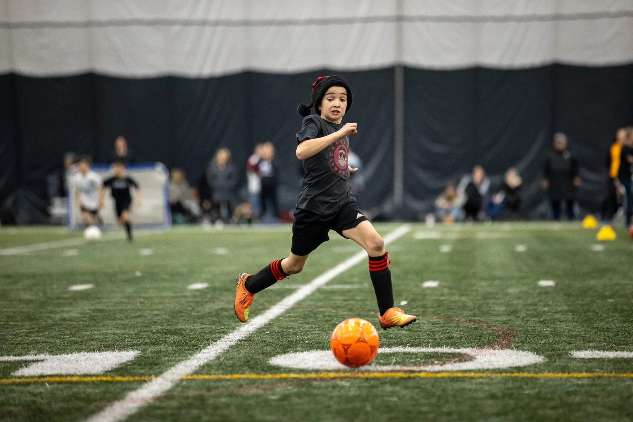 A young girl in sports gear running on an indoor soccer field with an orange soccer ball, with other kids and adults in the background.